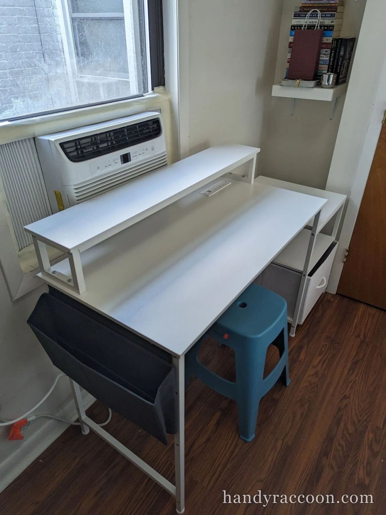White desk with a raised shelf assembled beside a window air conditioner and stool.