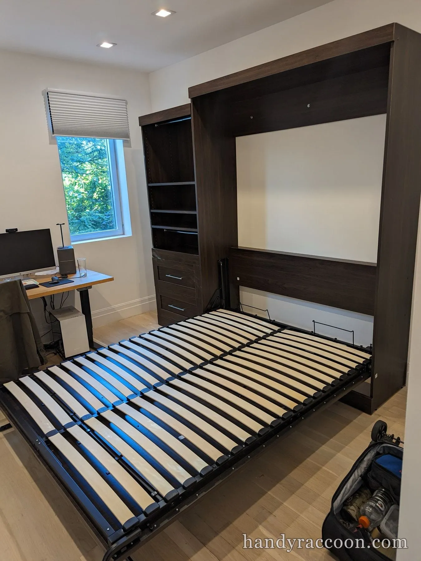 Dark wood Murphy wall bed folded down showing slats beside built-in shelves and a desk.