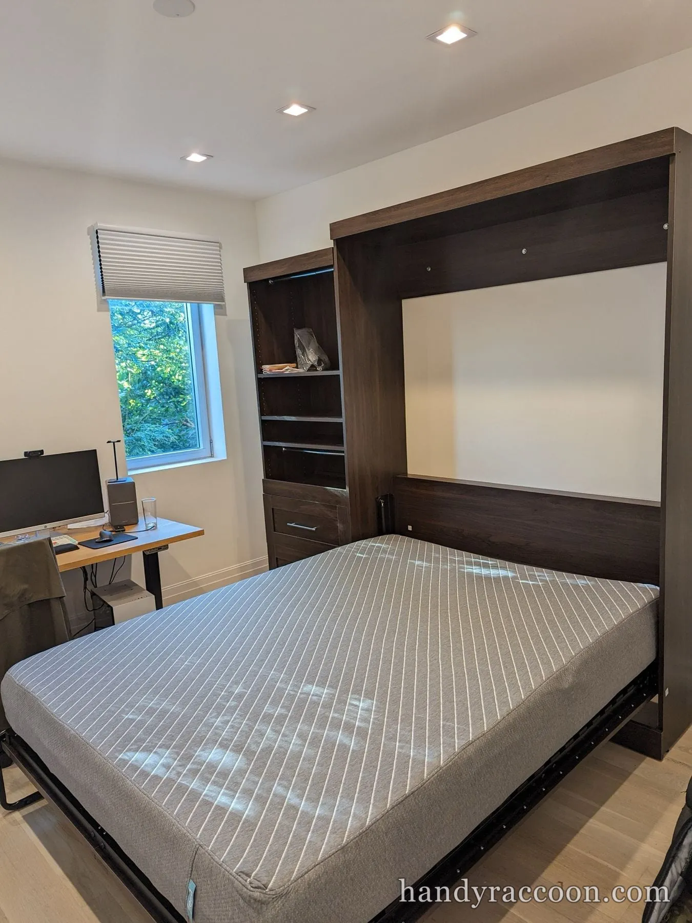 Dark wood Murphy bed pulled down showing a striped mattress, with a matching shelf unit to the left.