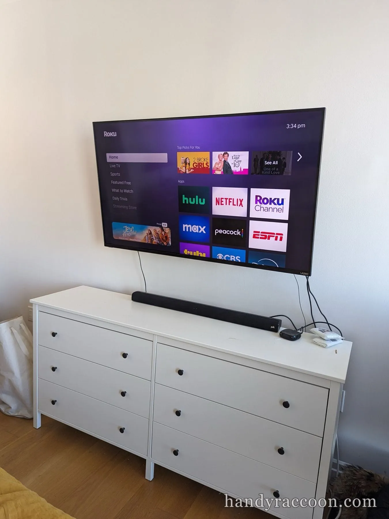 Flat-screen TV mounted on the wall with a soundbar over a white six-drawer dresser.