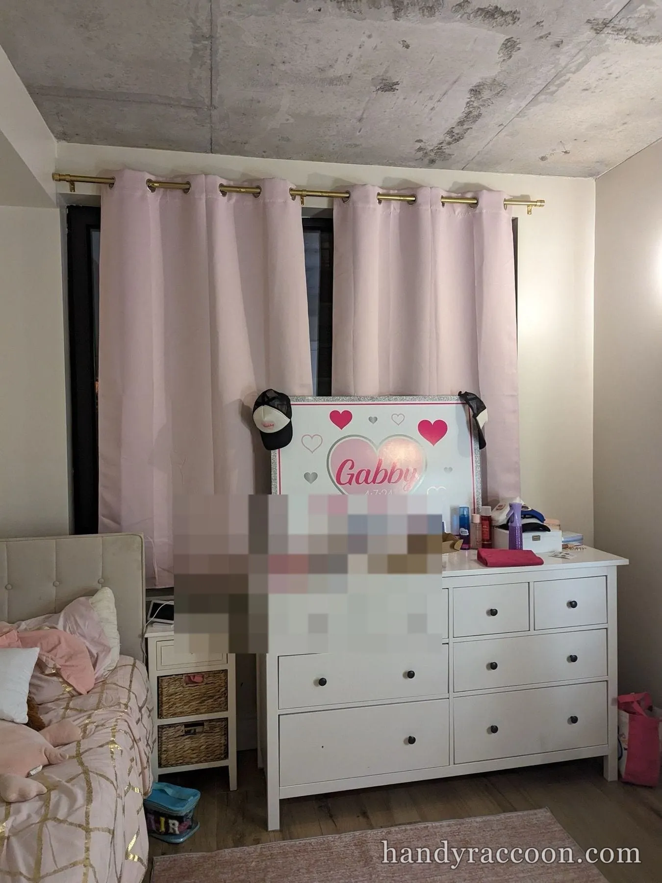 White eight-drawer dresser and hung pink curtains in a bedroom with a name sign and small photos.