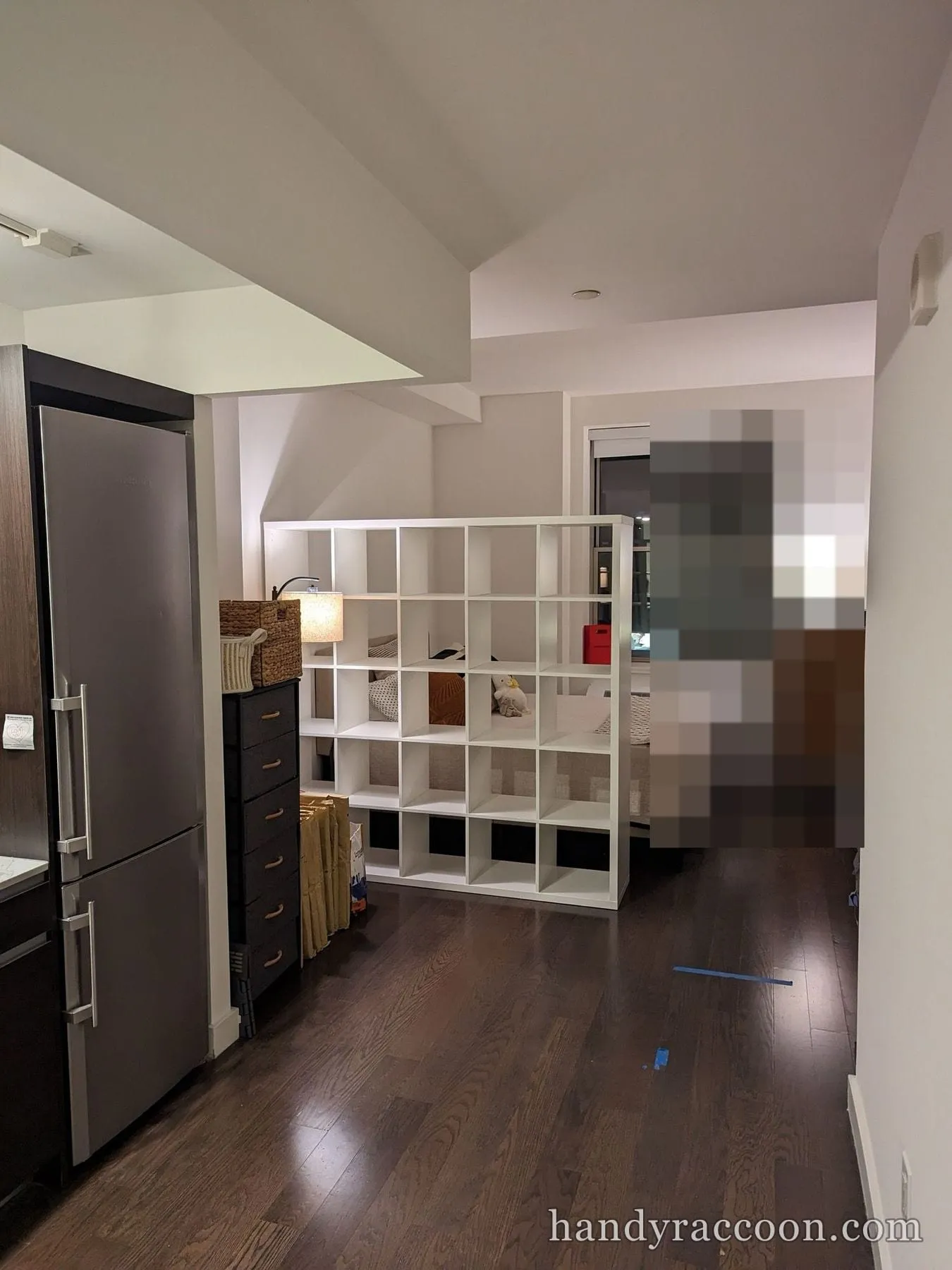 Tall white 5-by-5 cube shelving unit standing as a divider between a kitchen and a bed in a dim studio.