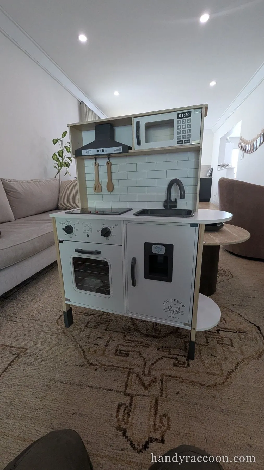 Fully assembled white and wood toy play kitchen with oven, sink, hood and microwave in a bright living room.
