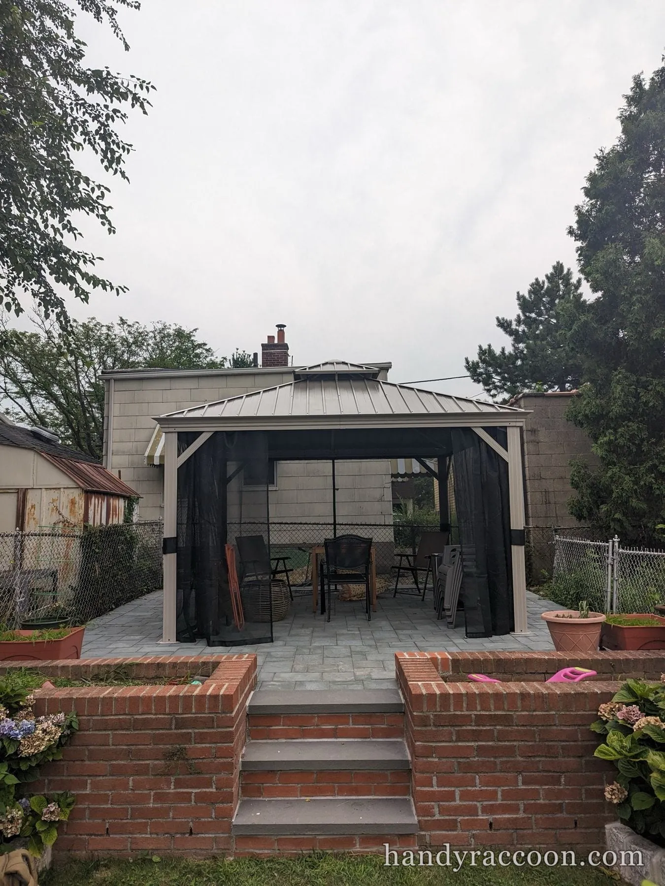 Assembled hardtop gazebo with dark mesh screen walls and patio chairs, brick steps in front.