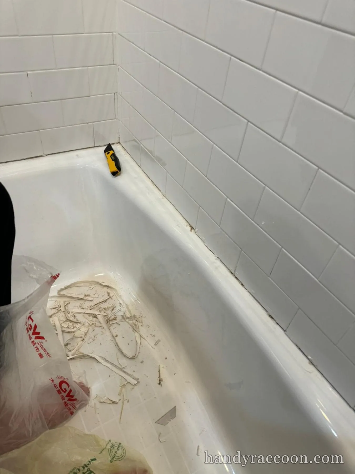 Utility knife and stripped-out old caulk in a white subway tile bathtub during a recaulk.