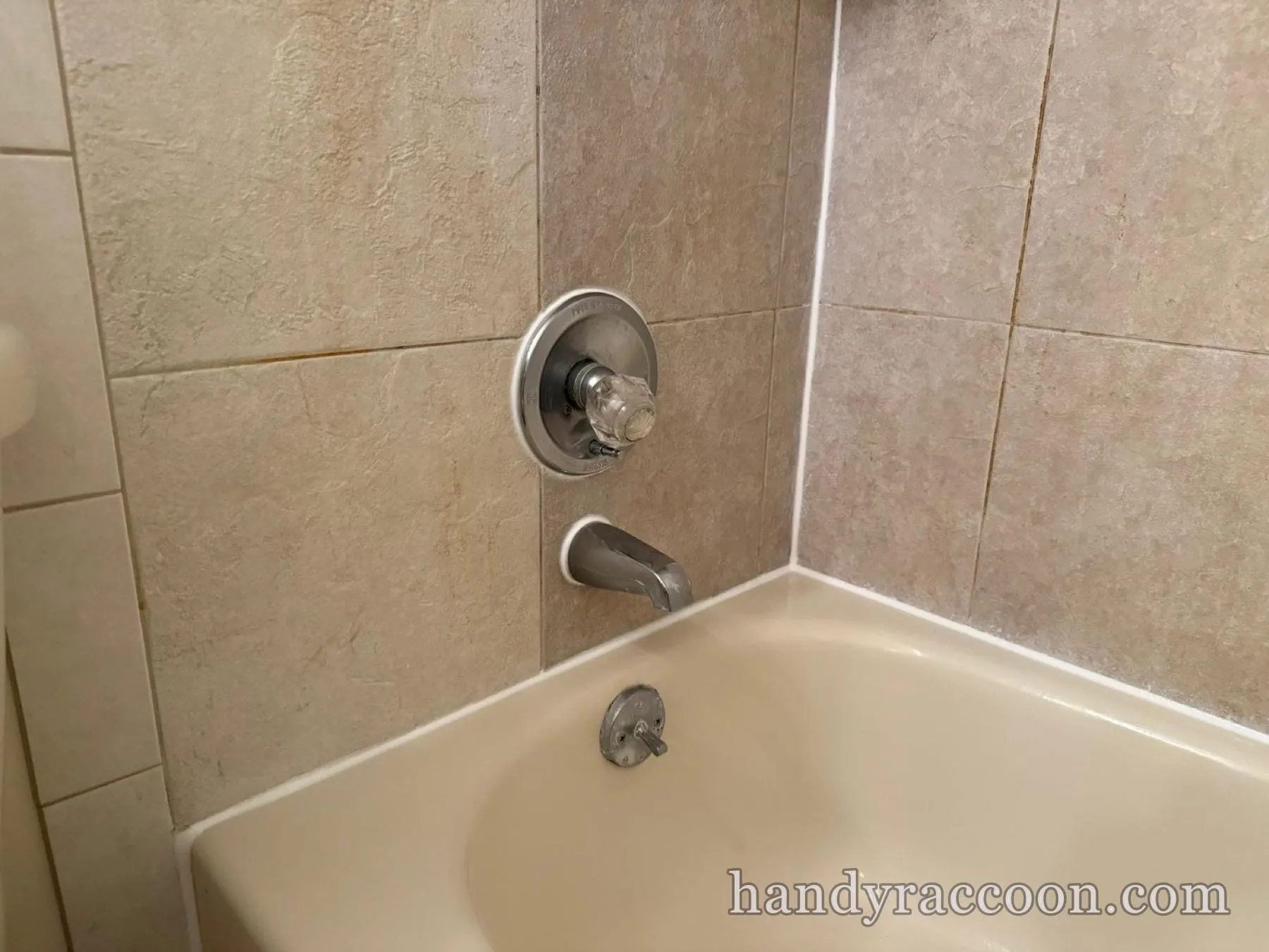 White caulk freshly applied around a chrome tub spout, valve and wall corner on beige tile.