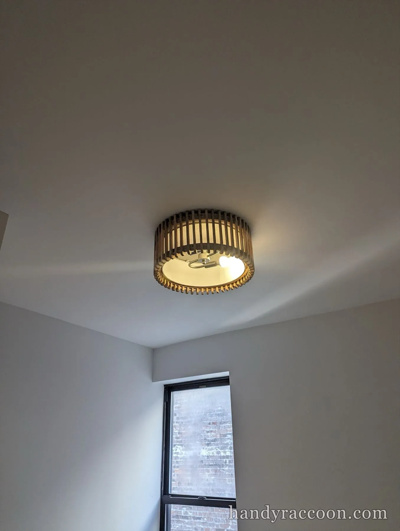 Round wood slat flush-mount ceiling light, lit, mounted on a white ceiling by a window.