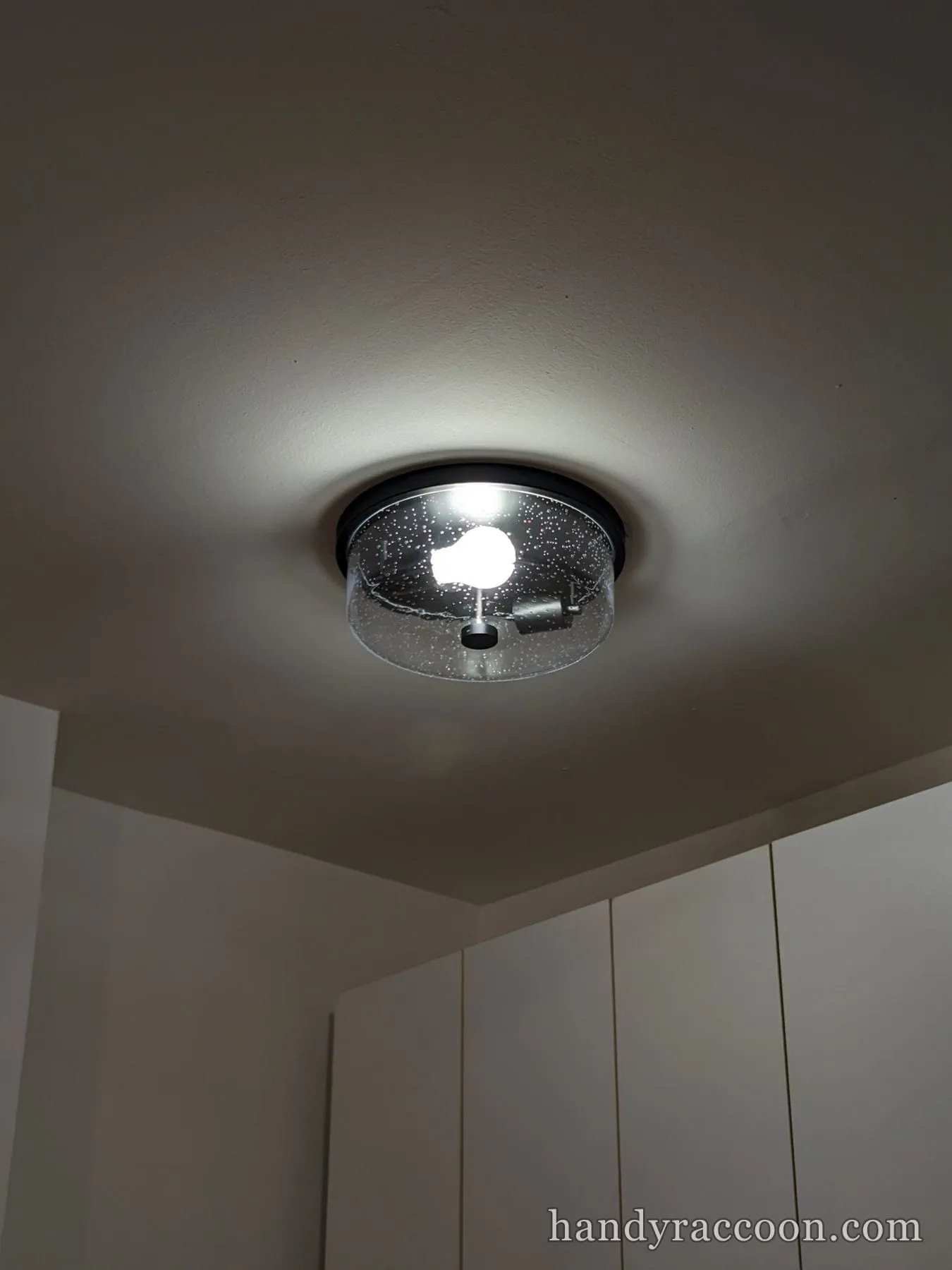 Clear seeded-glass drum flush-mount ceiling light lit in a room corner.