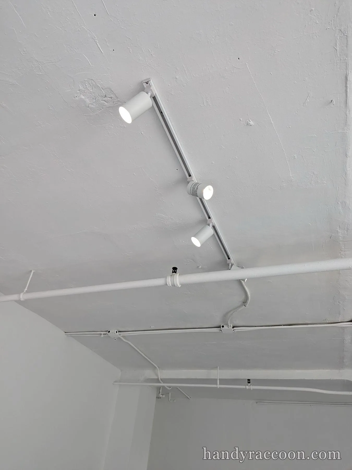 Three white cylinder track lights on a rail across an industrial white-painted loft ceiling.