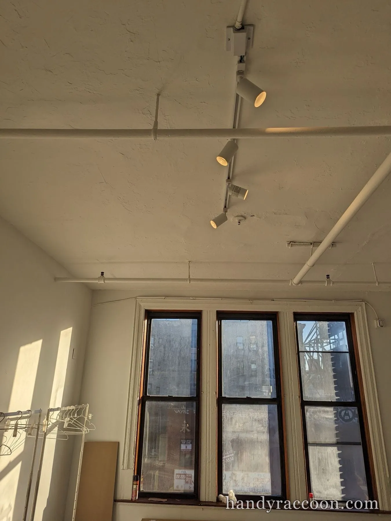 Loft room with track lighting on a pipe-lined ceiling, tall windows and a clothing rack below.
