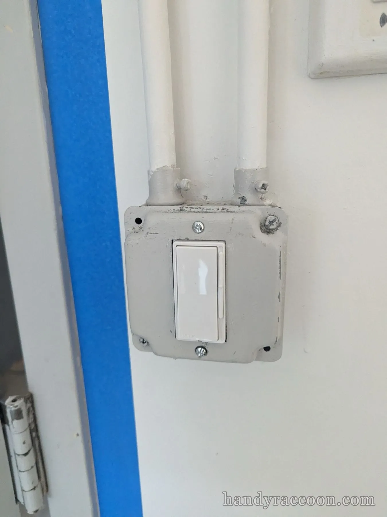 White rocker switch mounted in a gray metal surface box with two conduits, blue tape on a doorframe.