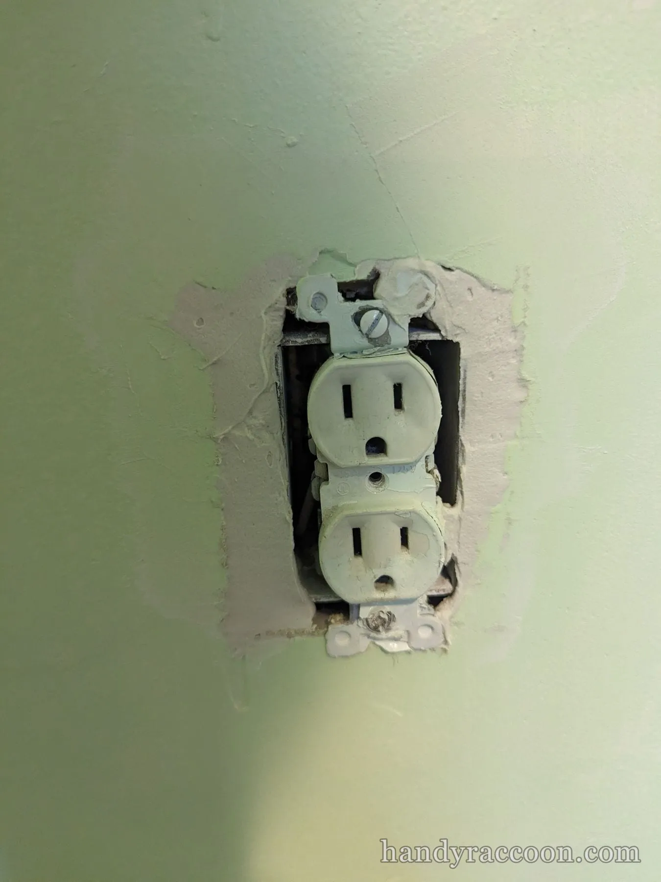 Old white duplex receptacle pulled from a wall box with chipped mint-green paint around it.
