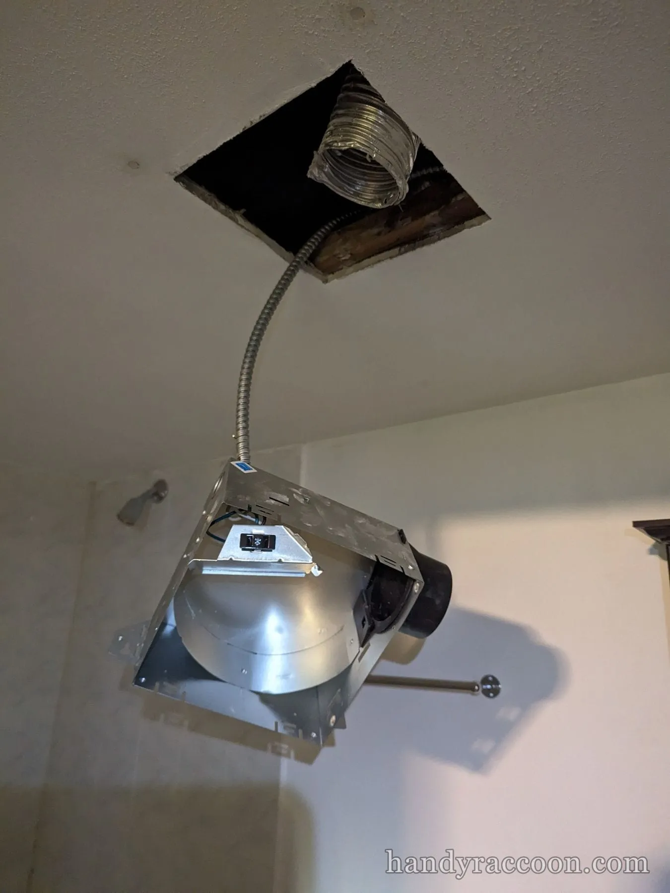 Metal bathroom exhaust fan housing hanging from a ceiling opening with flex duct and armored cable.