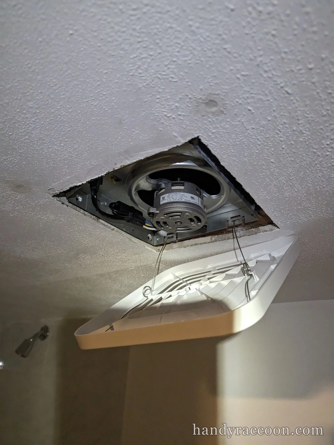 Bathroom exhaust fan housing in a popcorn ceiling with a white grille cover on spring clips.