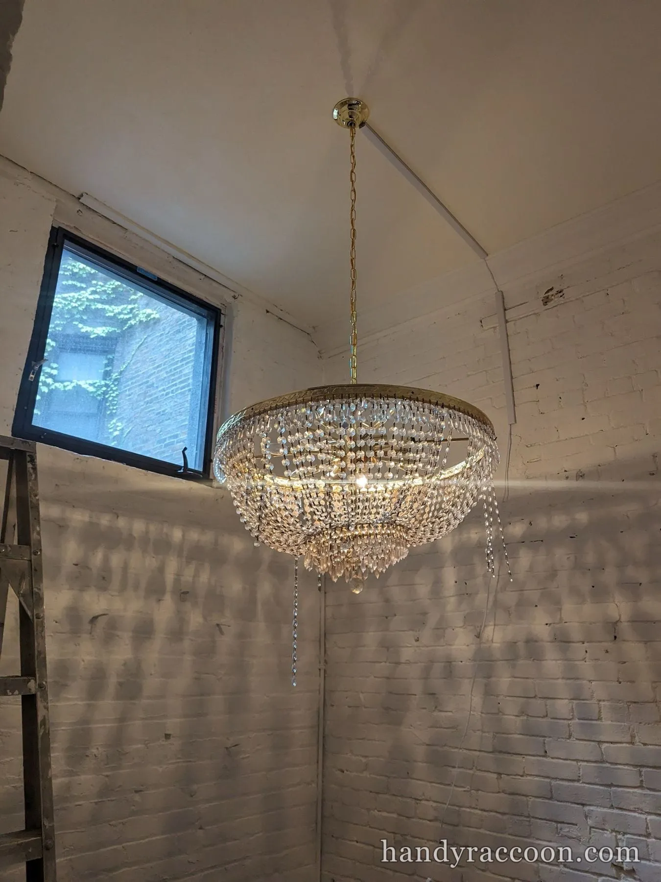Large gold and crystal chandelier installed and lit near a window in a brick-walled room.