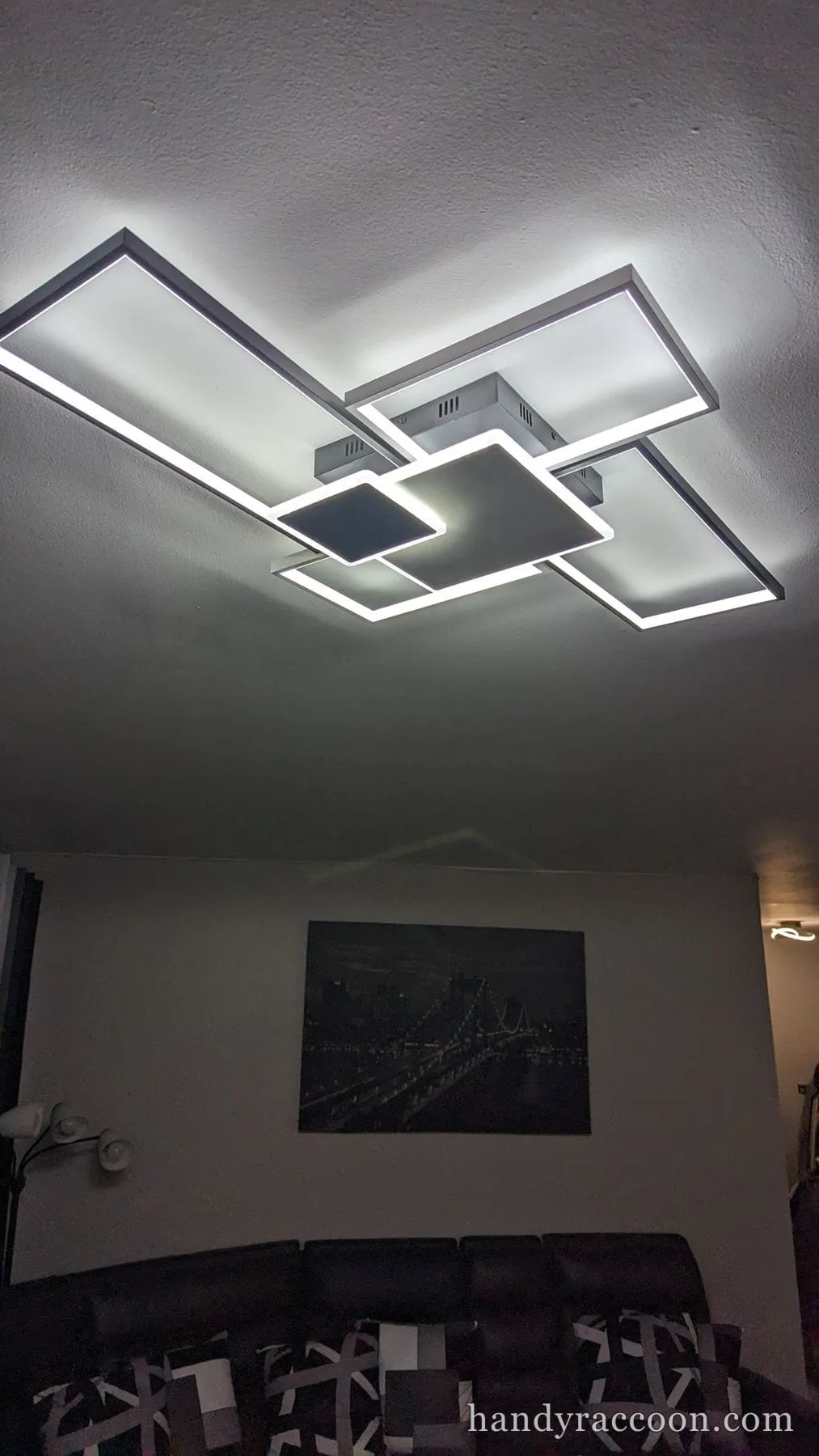 Modern overlapping-rectangle LED ceiling fixture glowing above a couch and wall art.