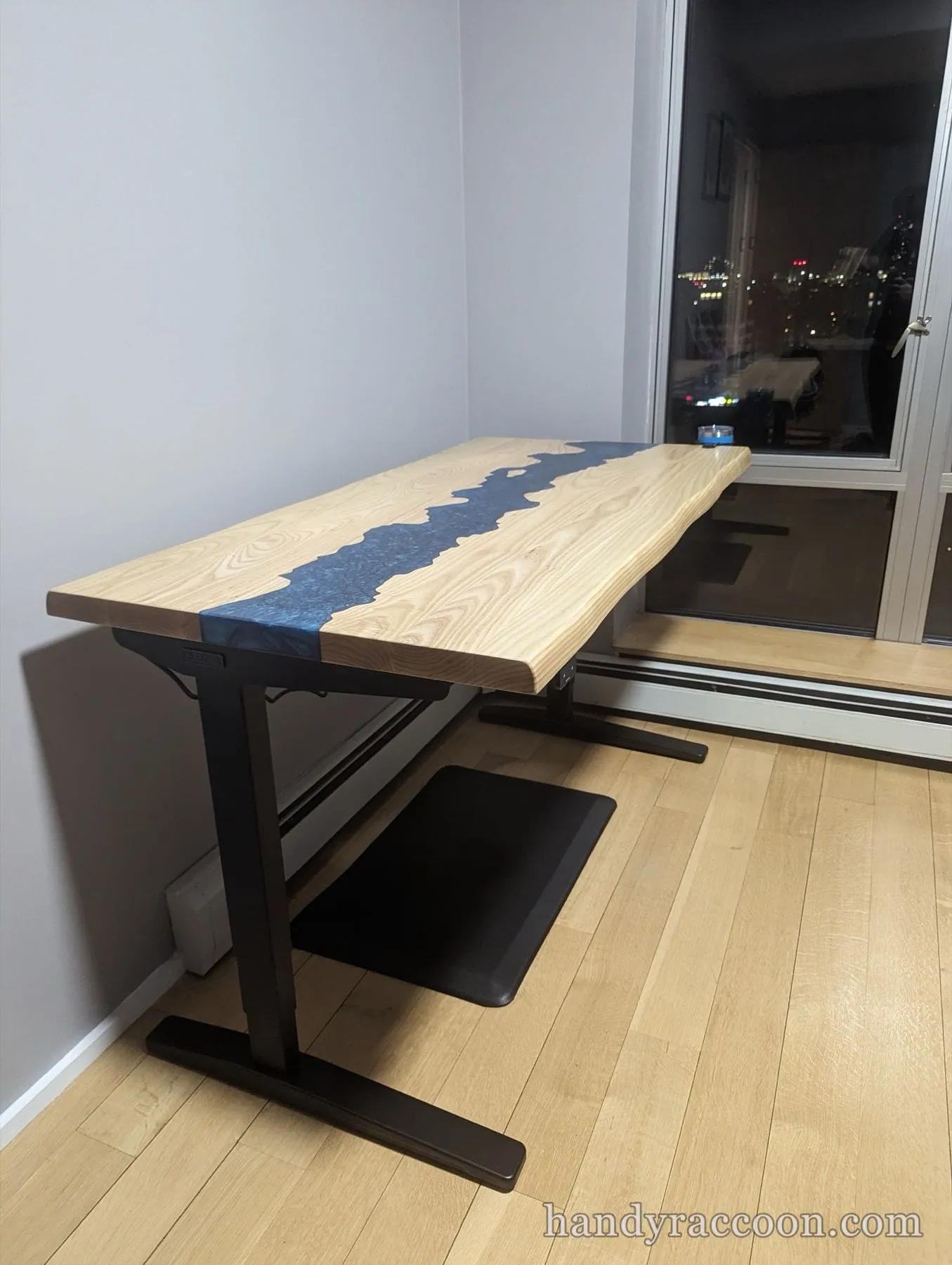 Live edge wood desk with a blue resin river top on black metal legs by a window.