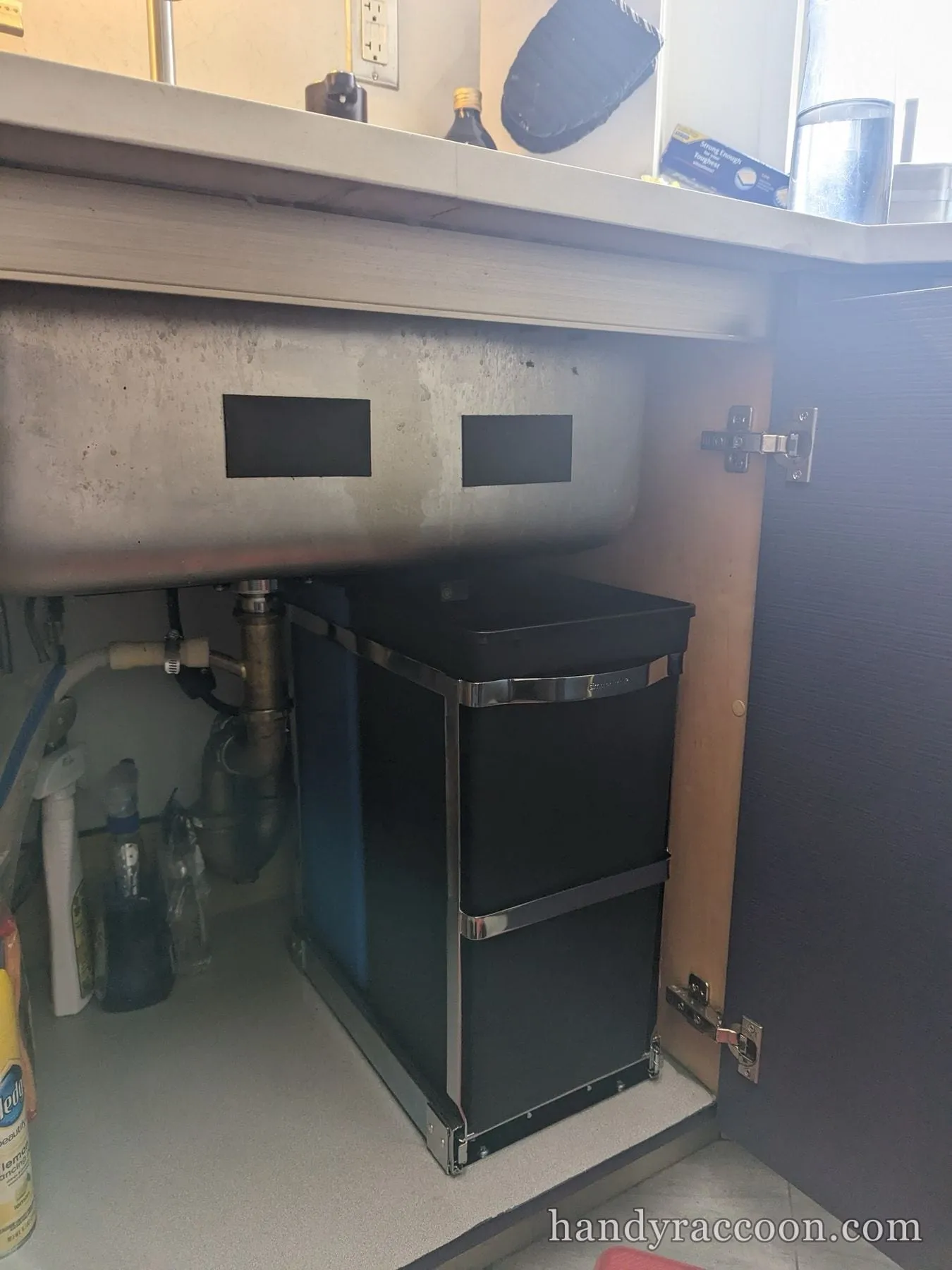 Black pull out waste bin on a chrome frame installed under a stainless kitchen sink.