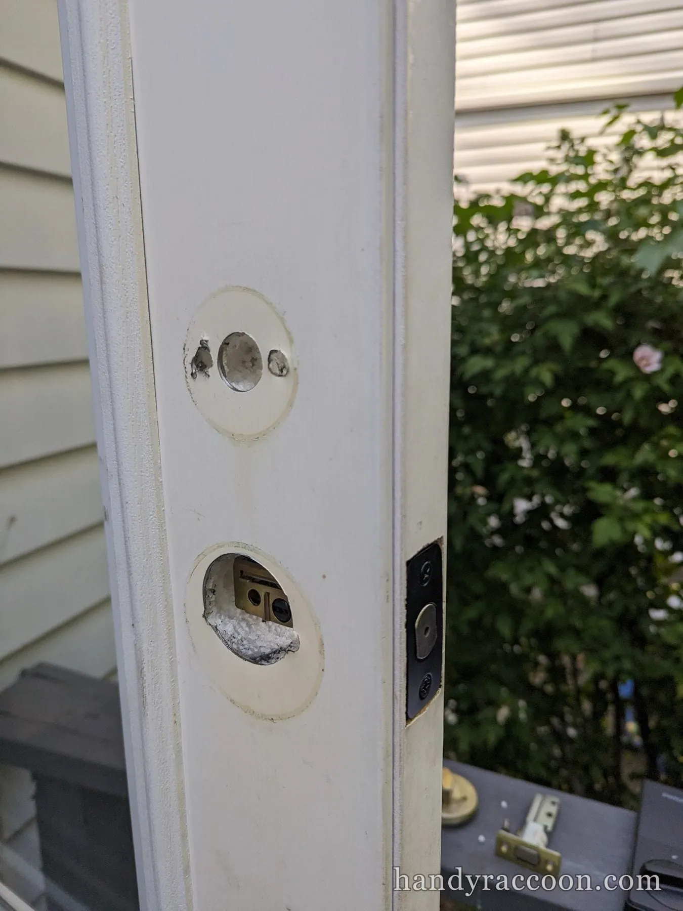 Edge of a white exterior door with two bored holes and a jamb strike mortise, old lock removed.
