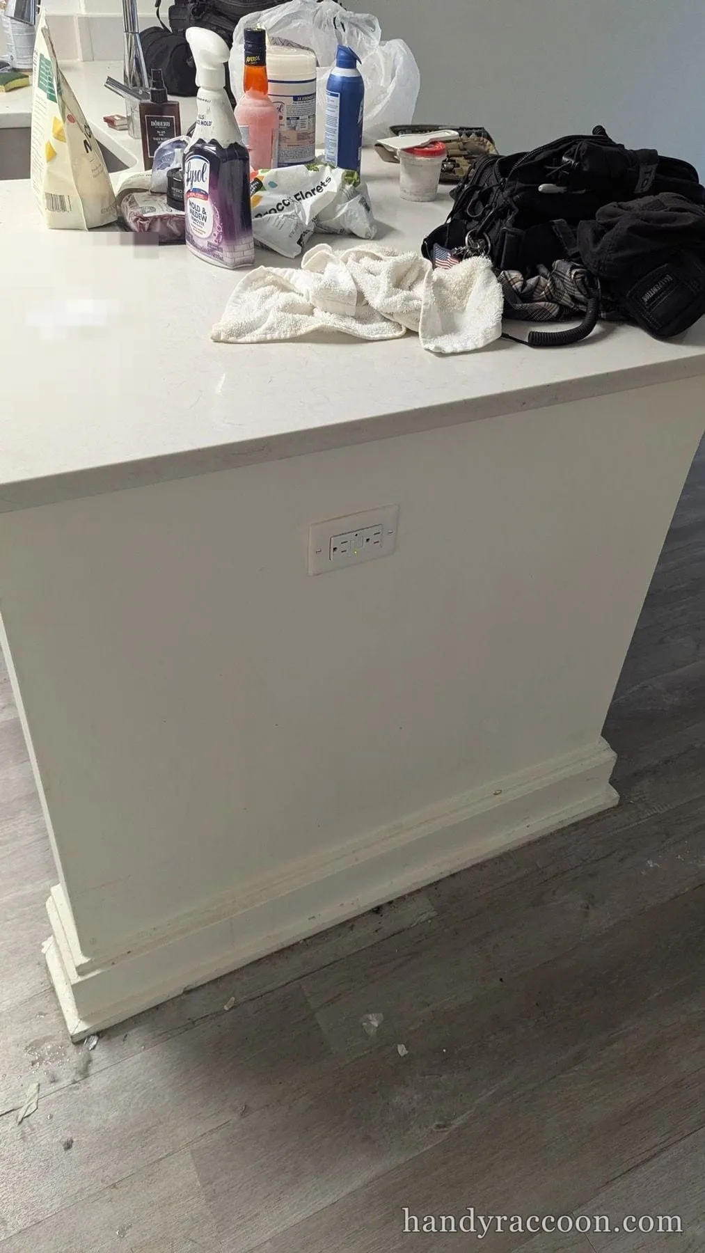 White kitchen island panel cleaned, with cluttered countertop of bottles, bags and towels above.