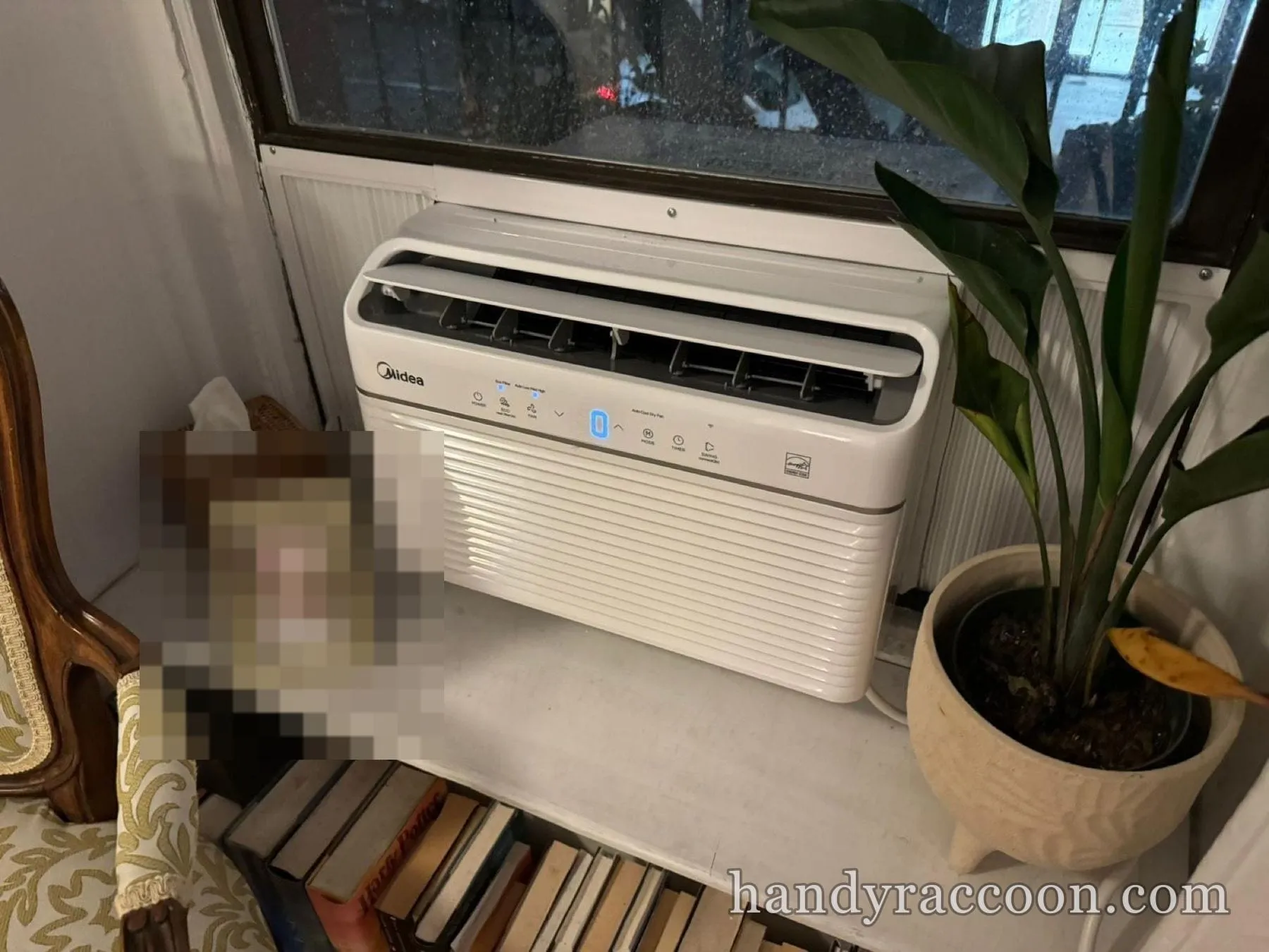 White Midea window air conditioner installed in a window above a shelf with books and a plant.