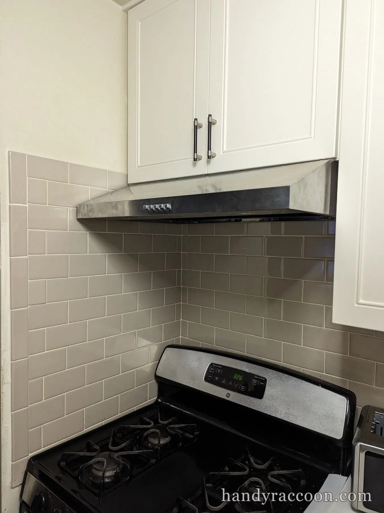Stainless steel range hood installed beneath white cabinets over a gas stove with subway tile.