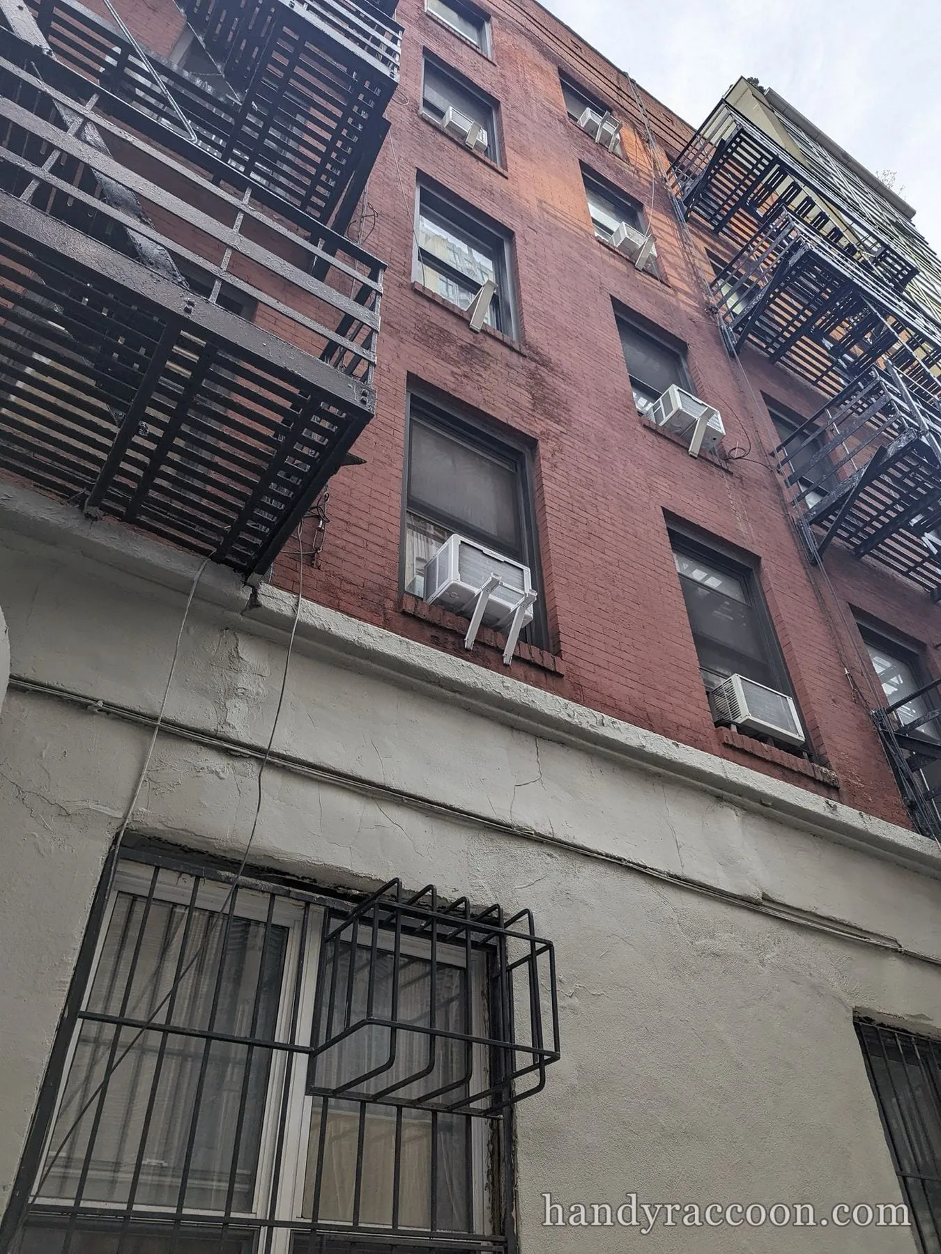 Brick apartment building facade with several window air conditioners on support brackets and fire escapes.