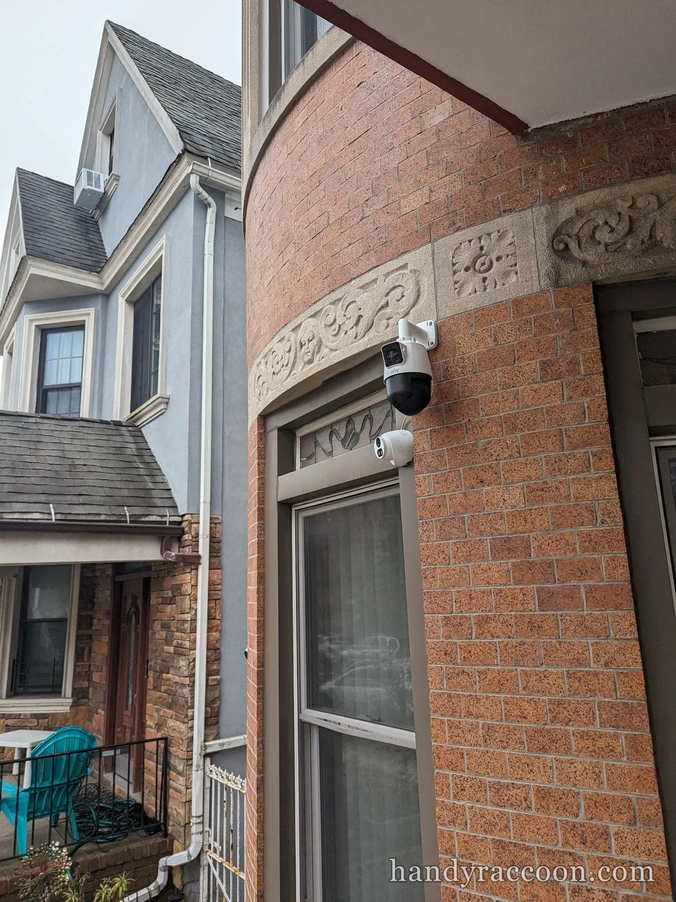 A white PTZ dome camera and a smaller bullet camera mounted on a brick building corner above a window, with neighboring houses in view.