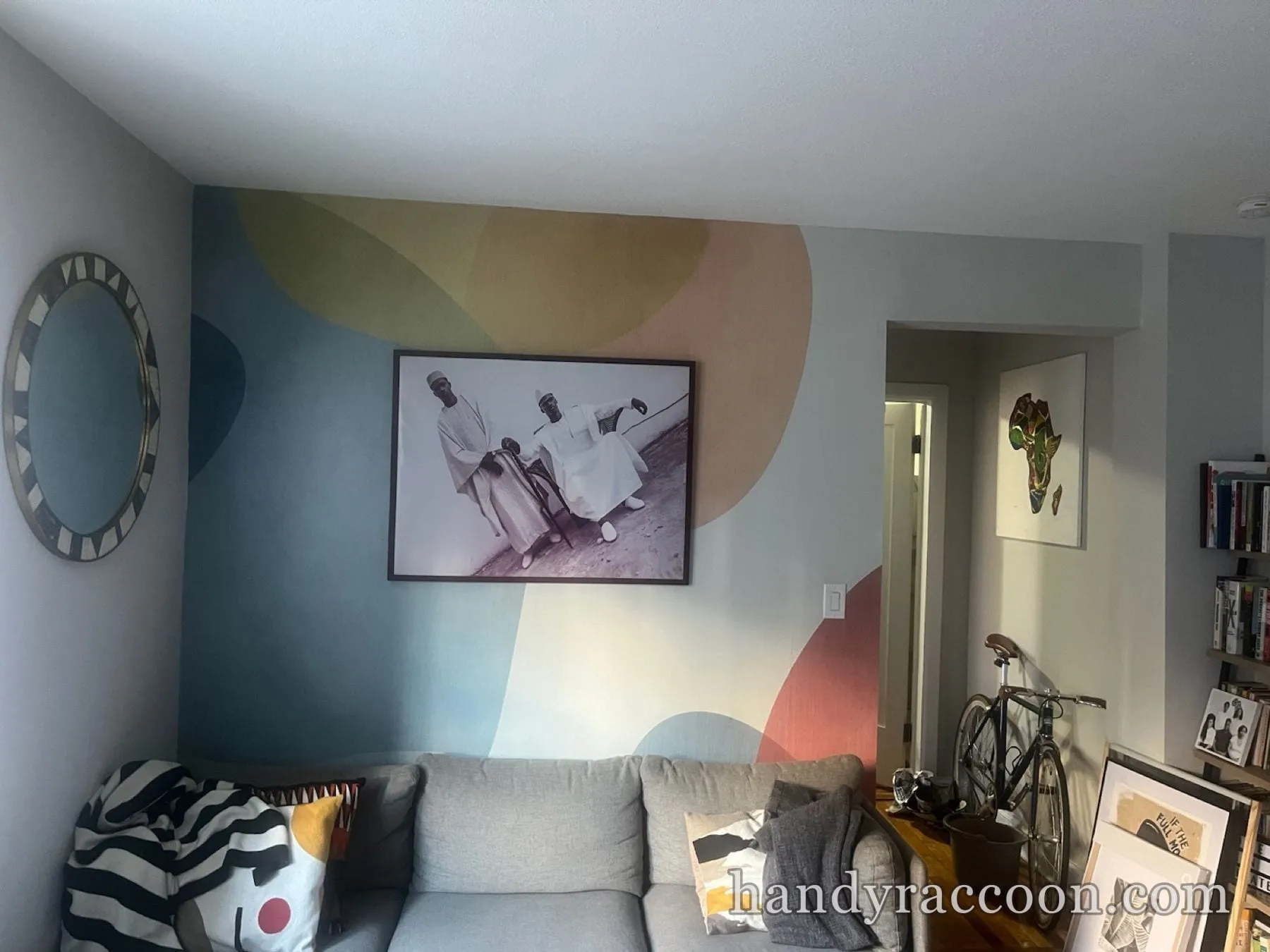 Living room with an accent wall of soft pastel color shapes, framed art, a round mirror, and a gray sofa.