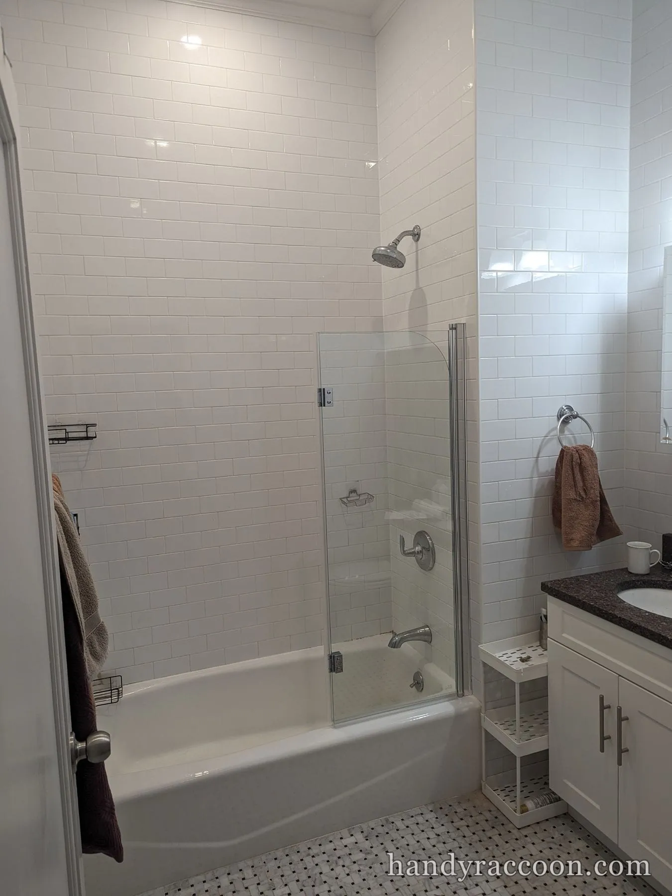 Hinged clear glass shower door on a white bathtub with white subway tile and a stone-top vanity.