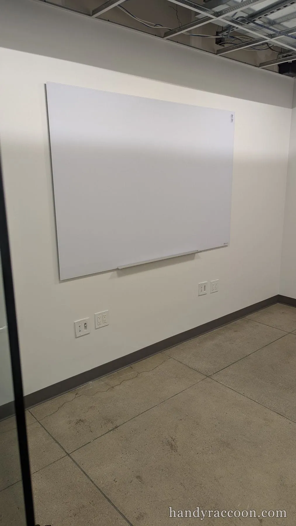 A white magnetic whiteboard mounted on a wall in an office with a concrete floor and exposed ceiling.