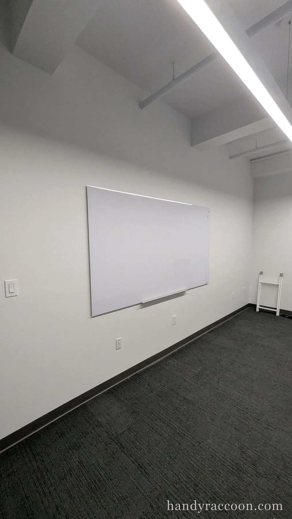 A white magnetic whiteboard mounted on a gray wall in an empty carpeted office.