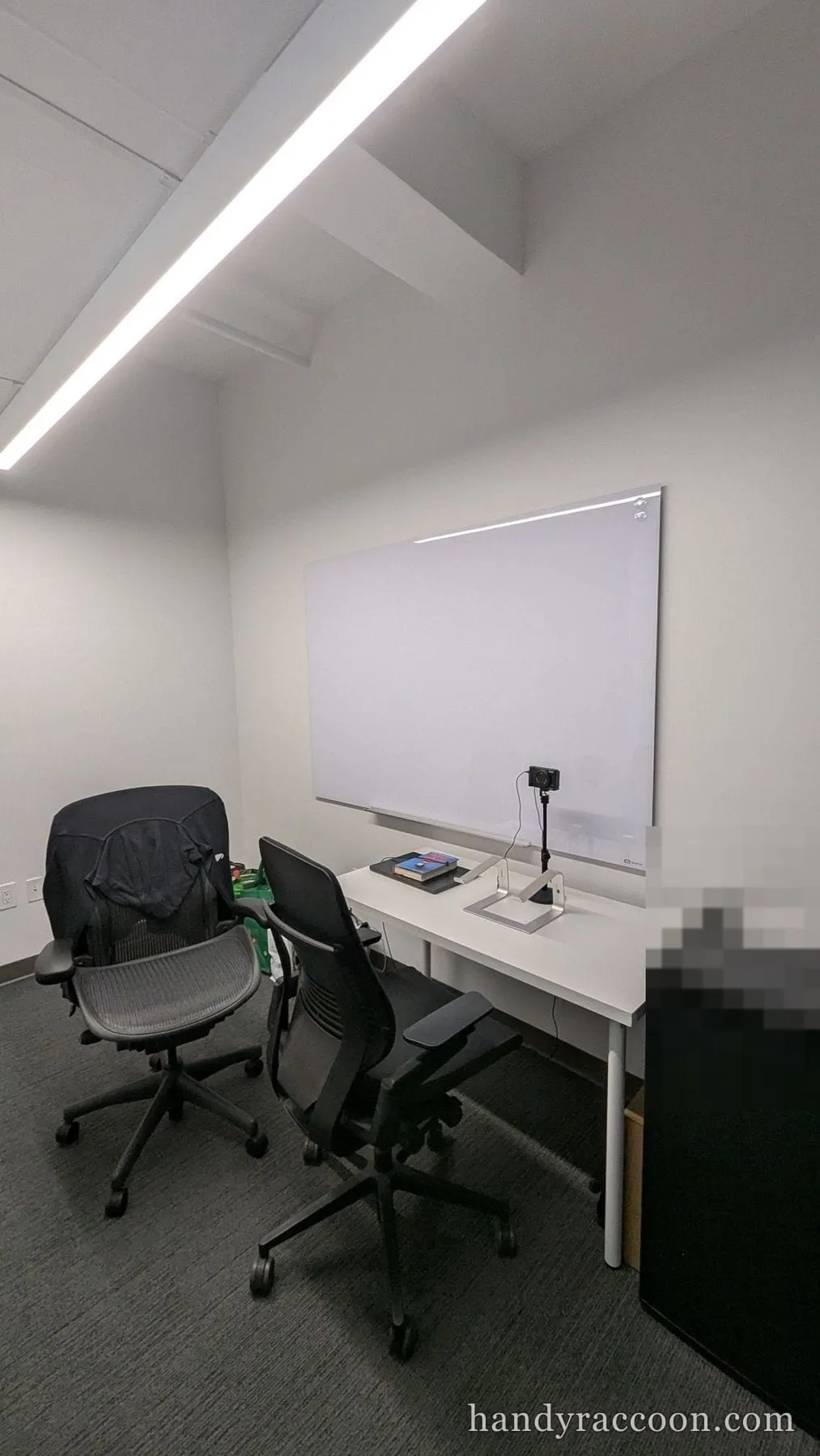 A glass dry-erase board mounted on a wall behind a desk with office chairs, a camera, and a mini fridge.