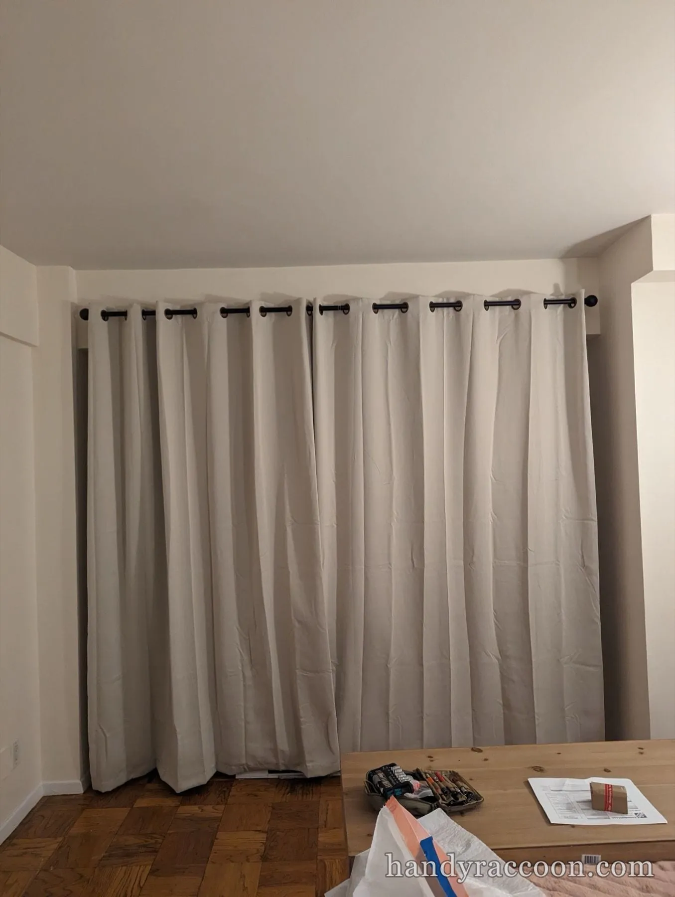 Beige blackout grommet curtains installed on a dark bronze curtain rod along a bedroom wall.