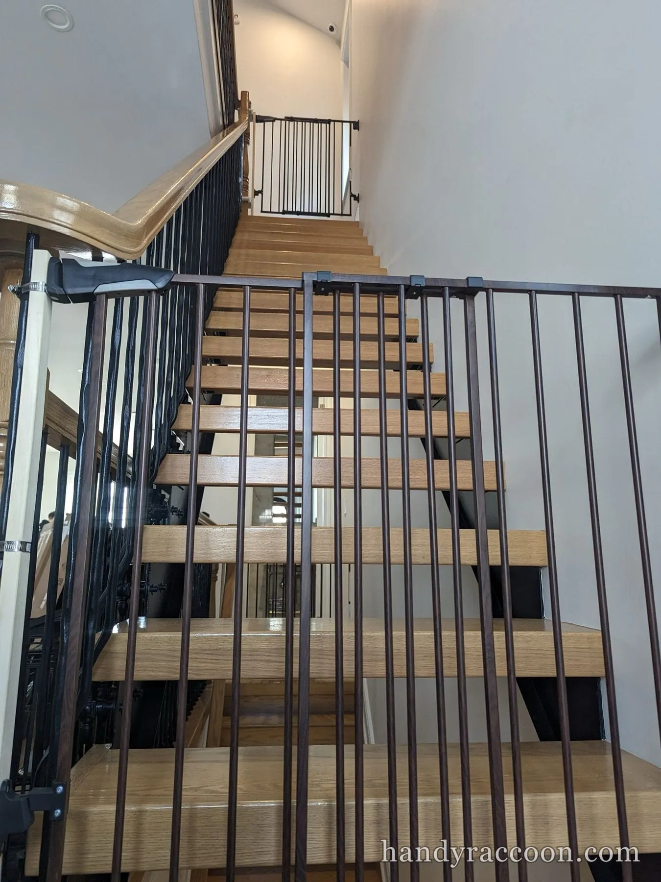 Dark metal safety gate mounted across a wooden open-tread staircase, white gate at the top landing