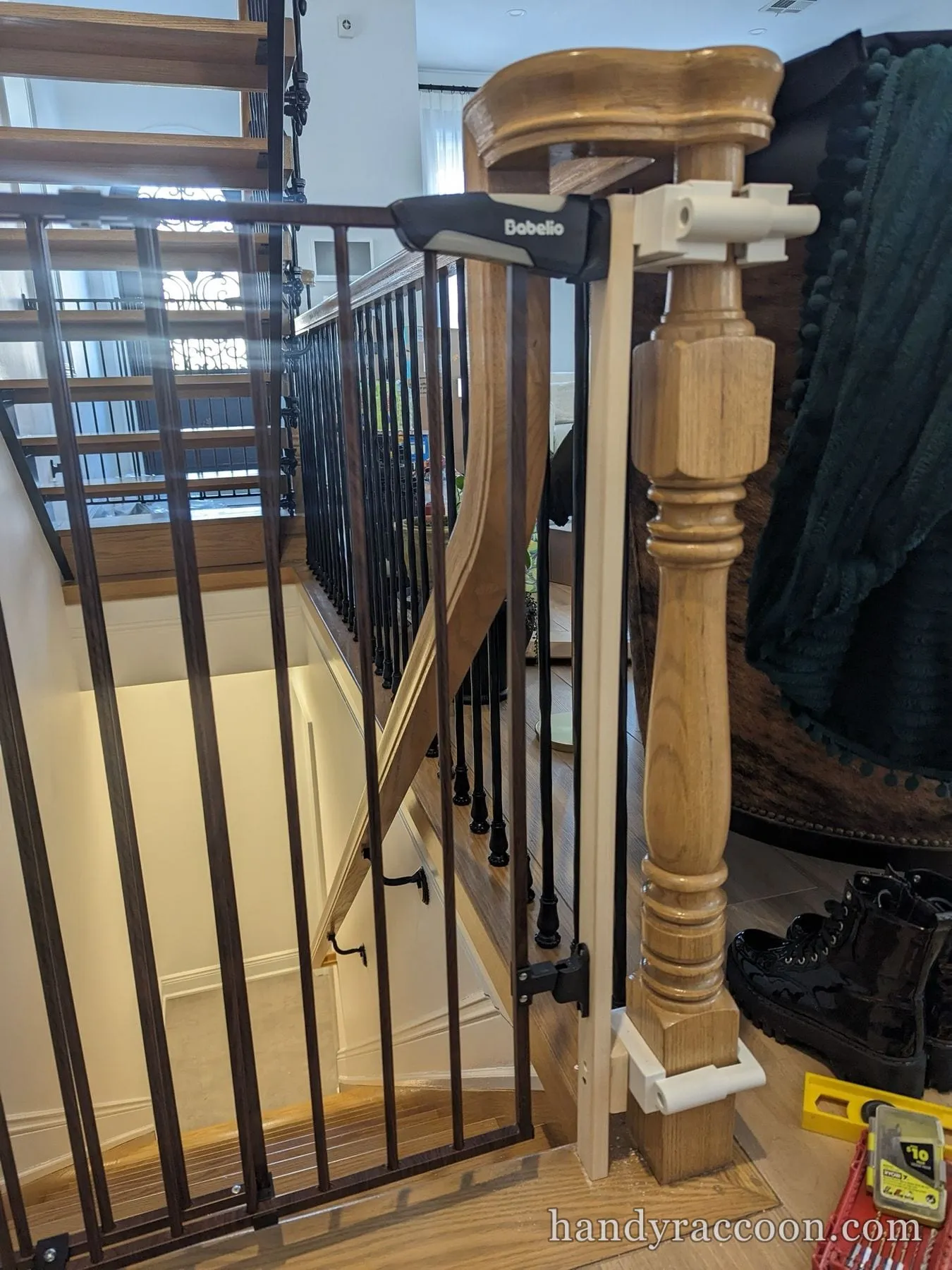 Baby safety gate clamped to a wooden newel post at the top of a staircase, boots and tools on the floor