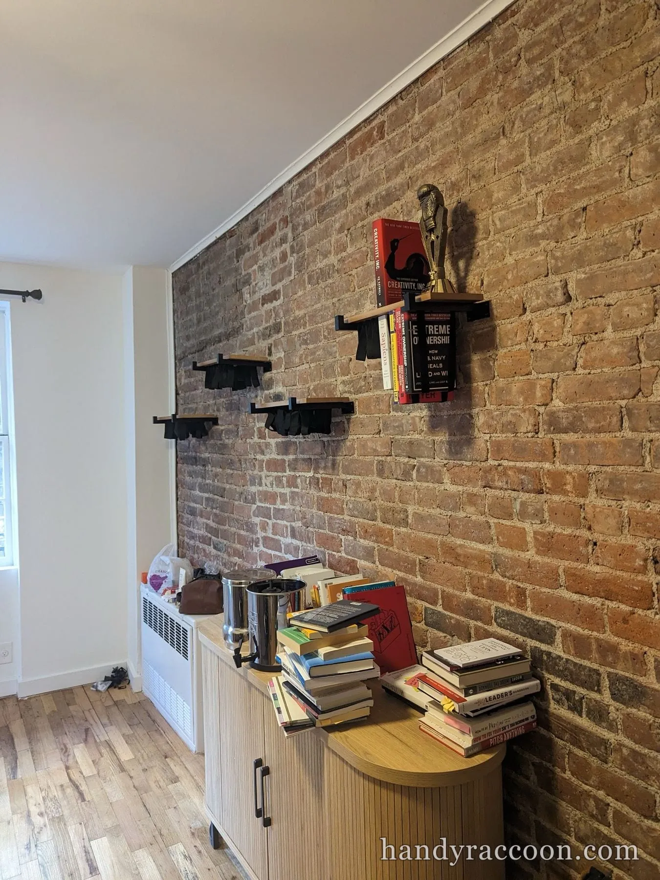 Exposed brick wall with several staggered wood floating shelves on black brackets holding books, cabinet below.