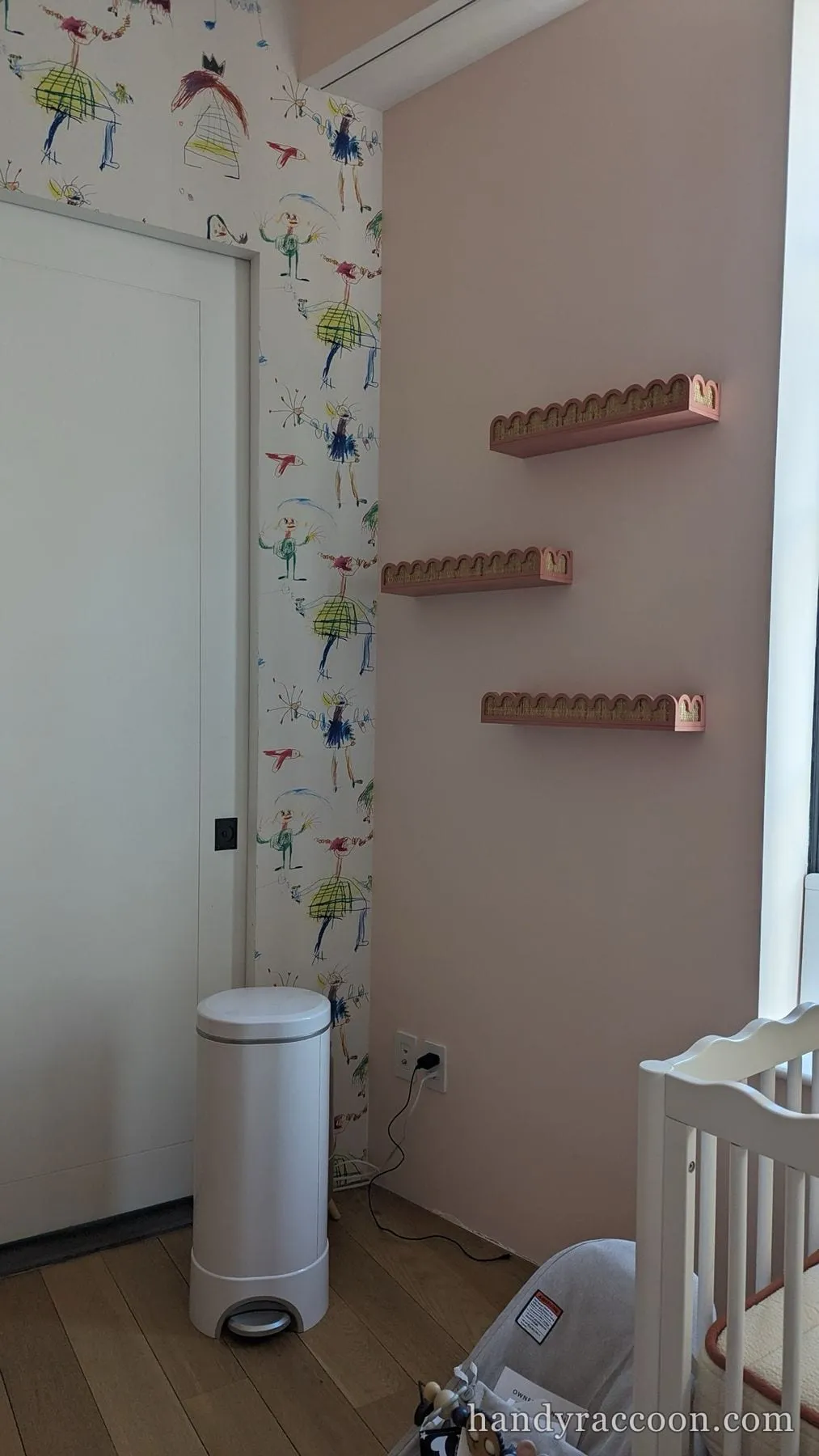 Nursery corner with pink wall and three pink scalloped shelves mounted in a staggered line near a white crib.