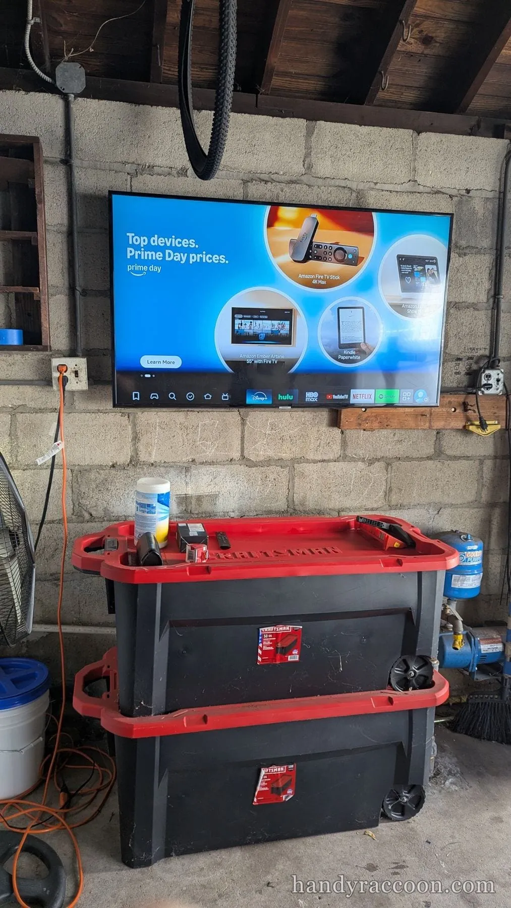 Flat-screen TV mounted on a cinderblock garage wall above red storage bins and a fan.
