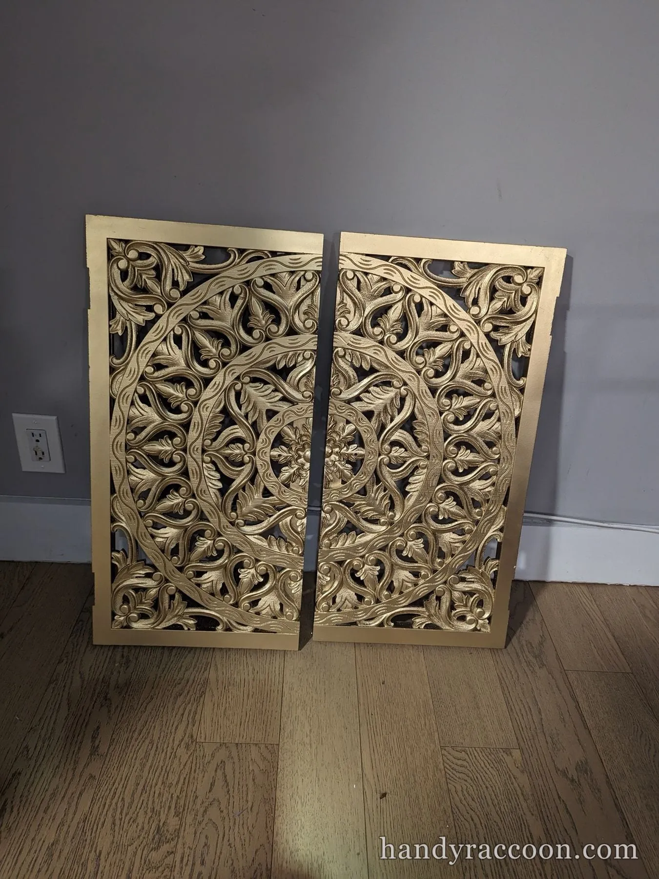 Two matching ornate carved openwork panels painted gold, leaning against a grey wall.