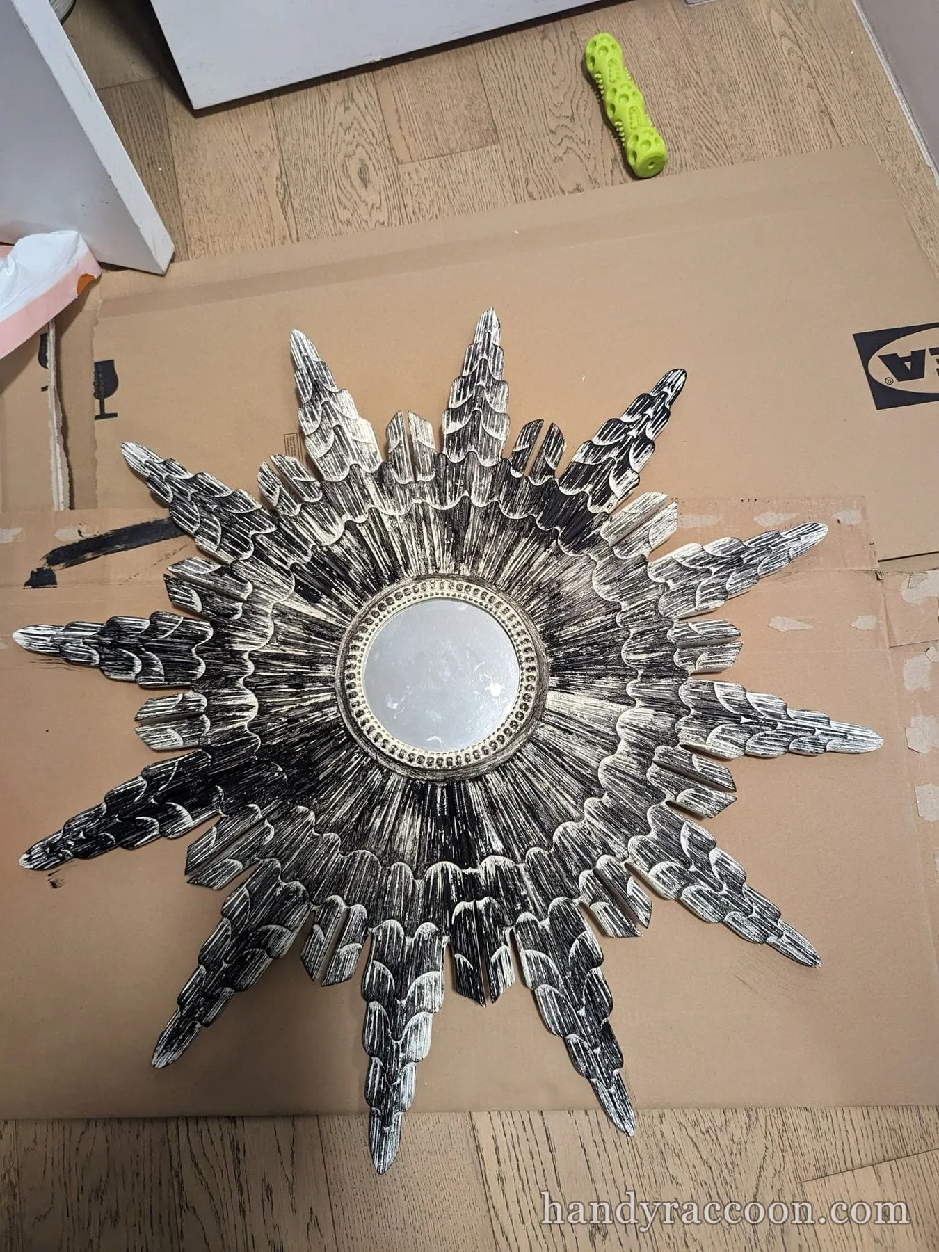 Ivory sunburst wall mirror with layered feathered rays lying on cardboard on the floor.