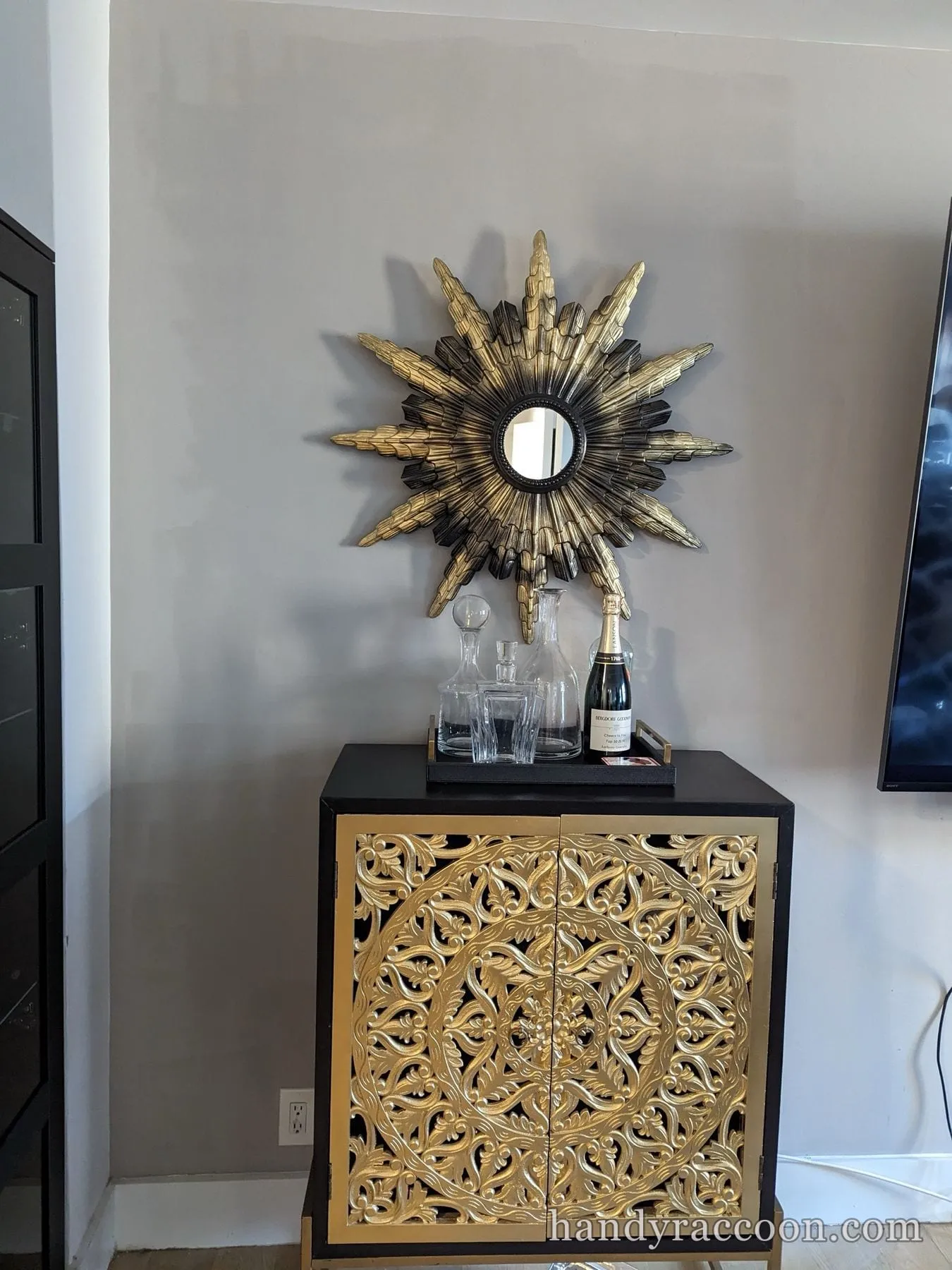 Gold and black sunburst wall mirror mounted above a black cabinet topped with glass decanters.