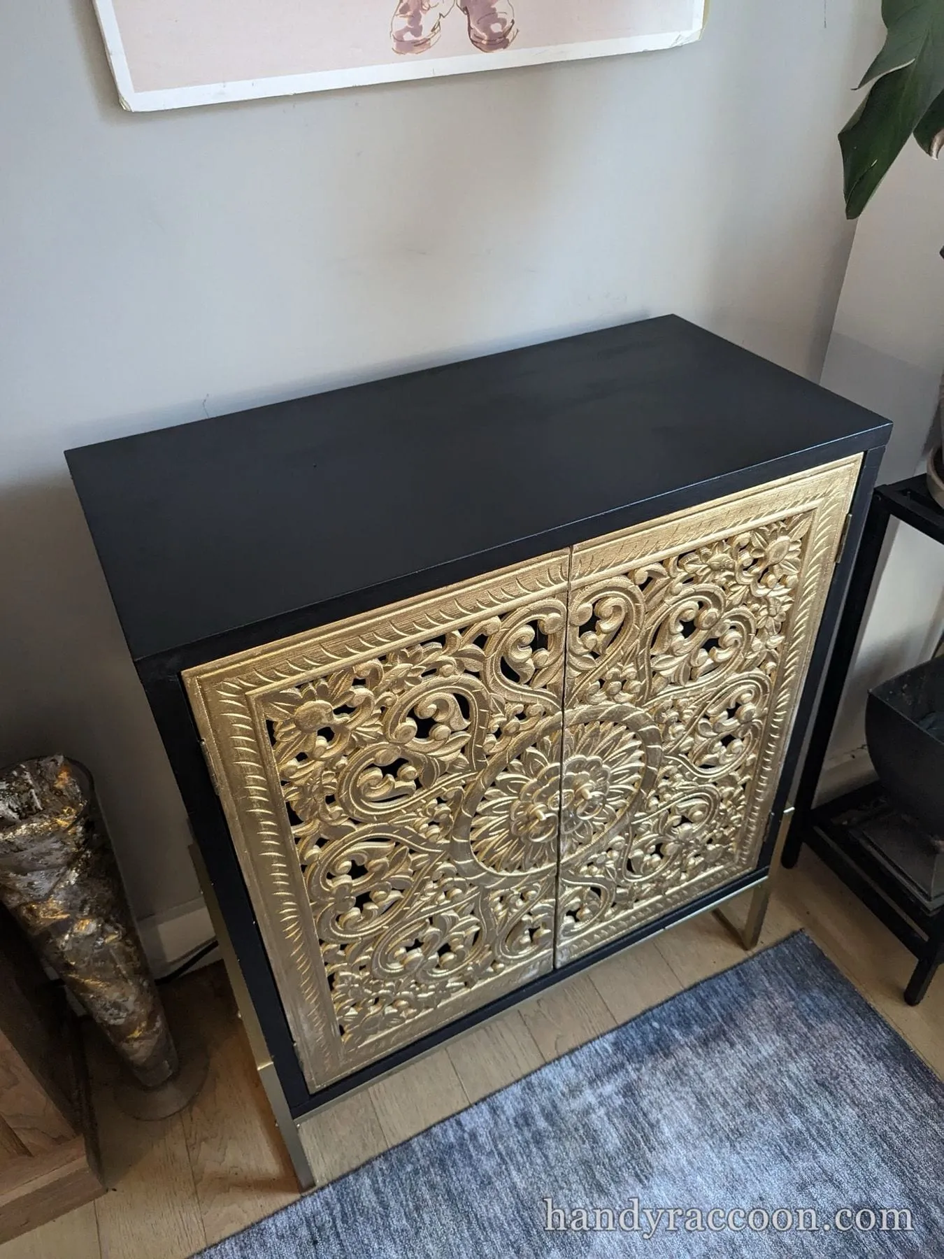 Black cabinet with ornate gold openwork carved doors on slim gold legs against a light wall.