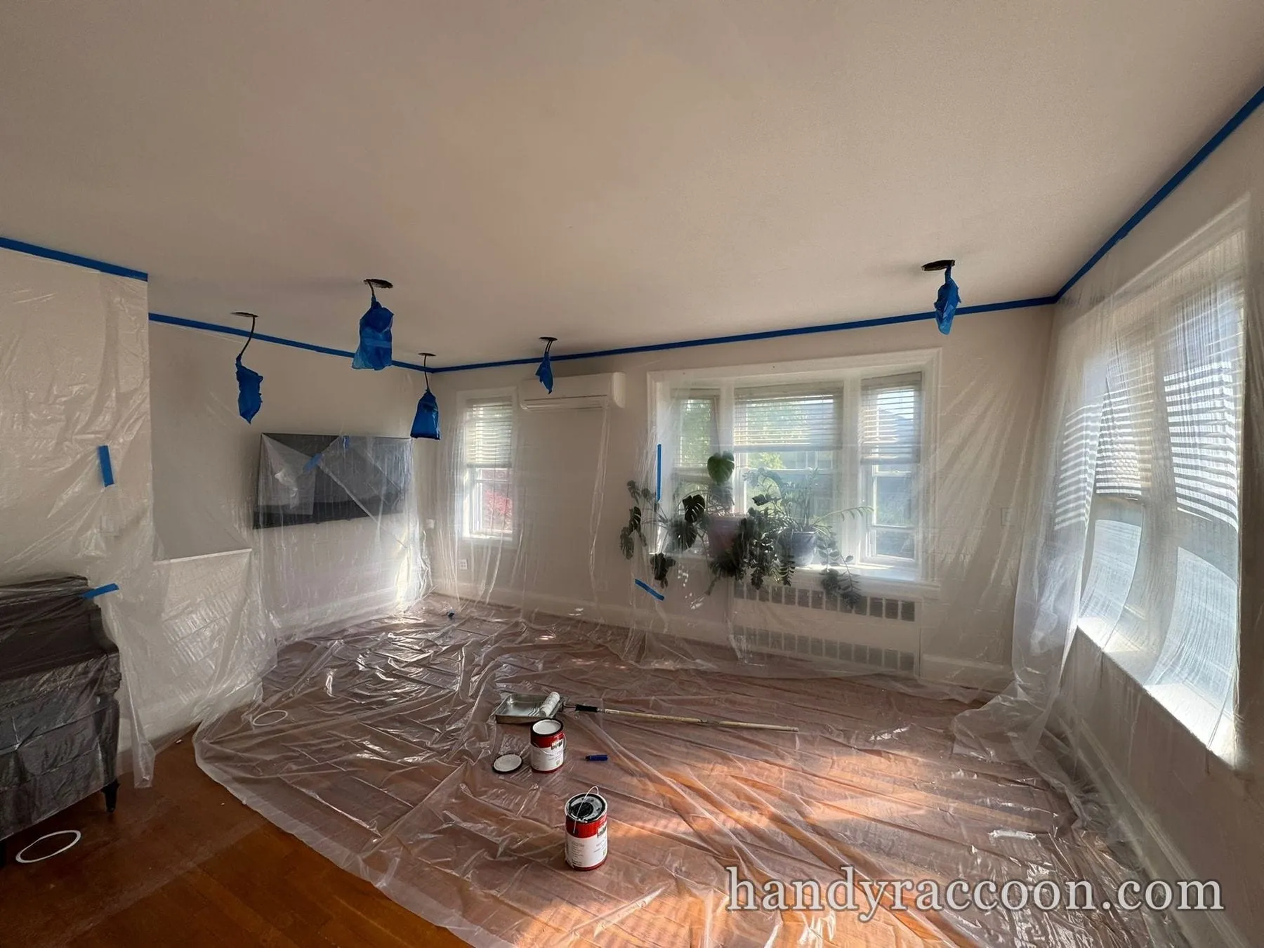Living room with floor, bay windows, and furniture covered in plastic sheeting and blue tape.
