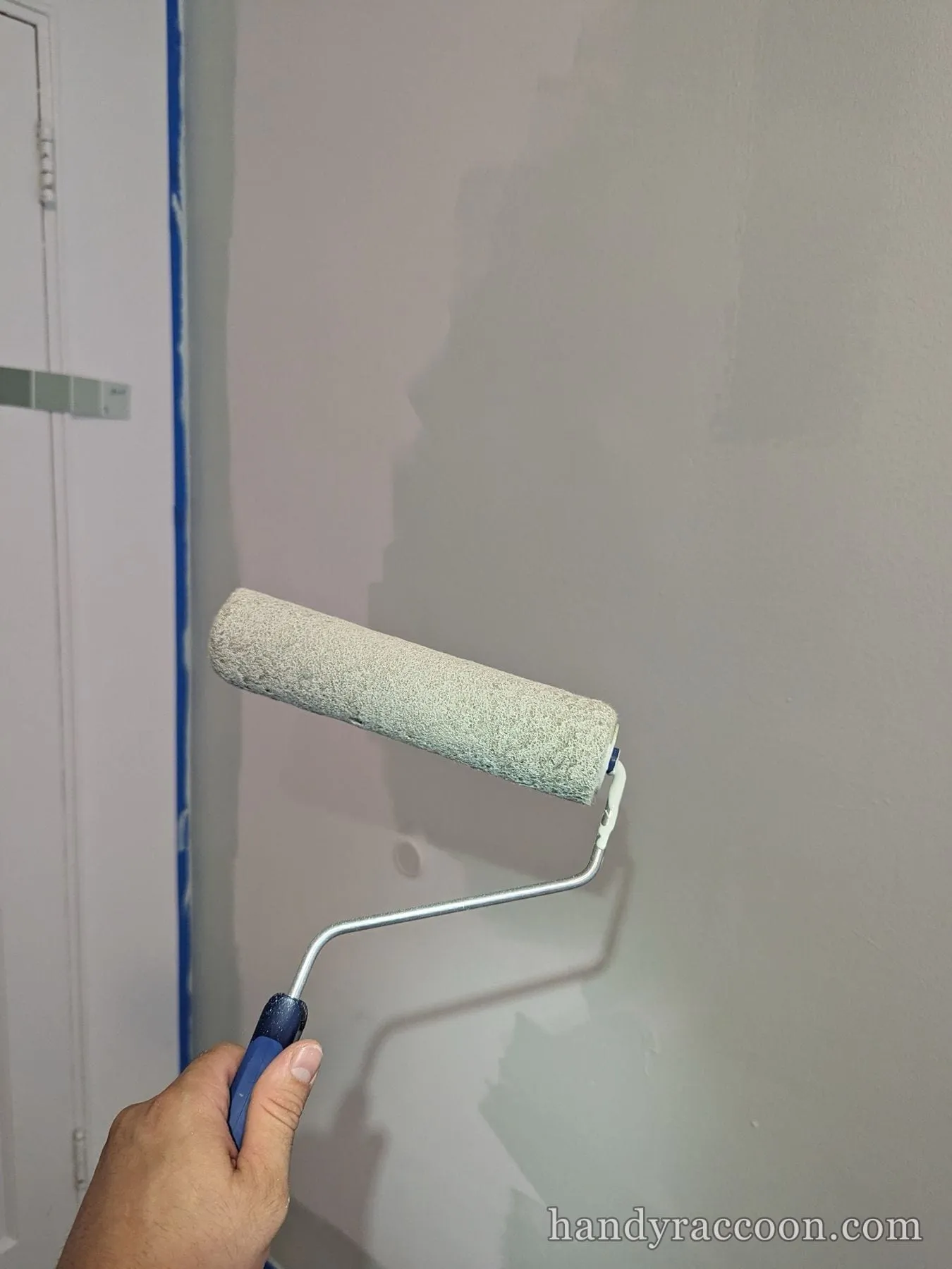 Hand holding a paint roller applying paint to a wall next to a blue-taped door frame.