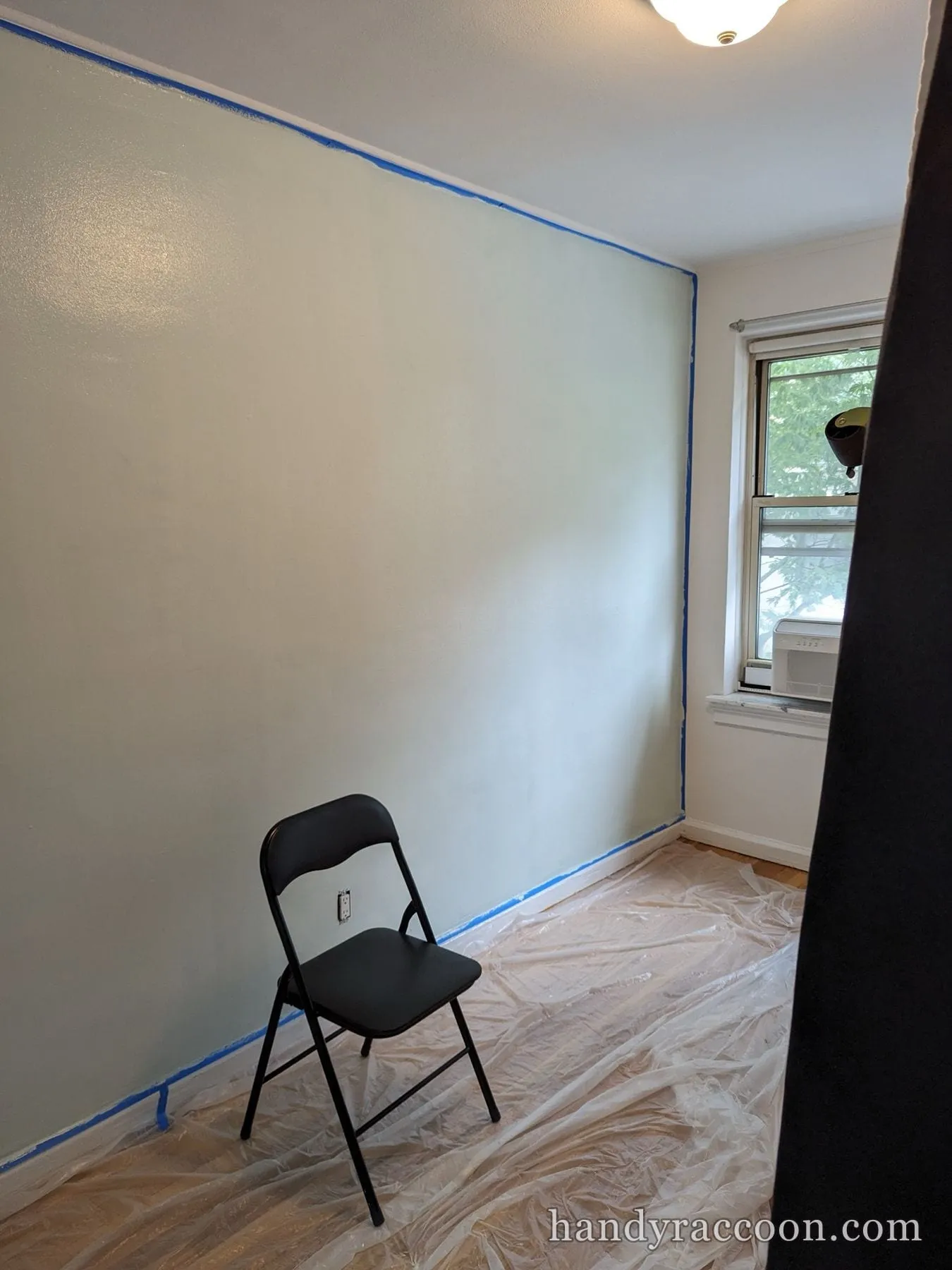 Empty room with a freshly painted pale sage-green wall, folding chair, and plastic floor cover.