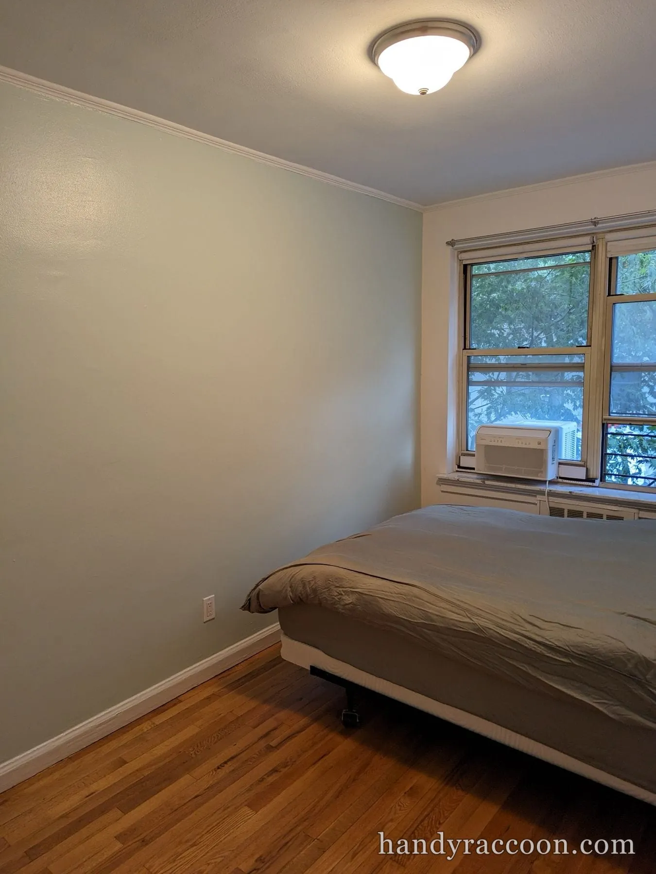 Furnished bedroom with soft sage-green walls, white trim, hardwood floor, and a window AC unit.