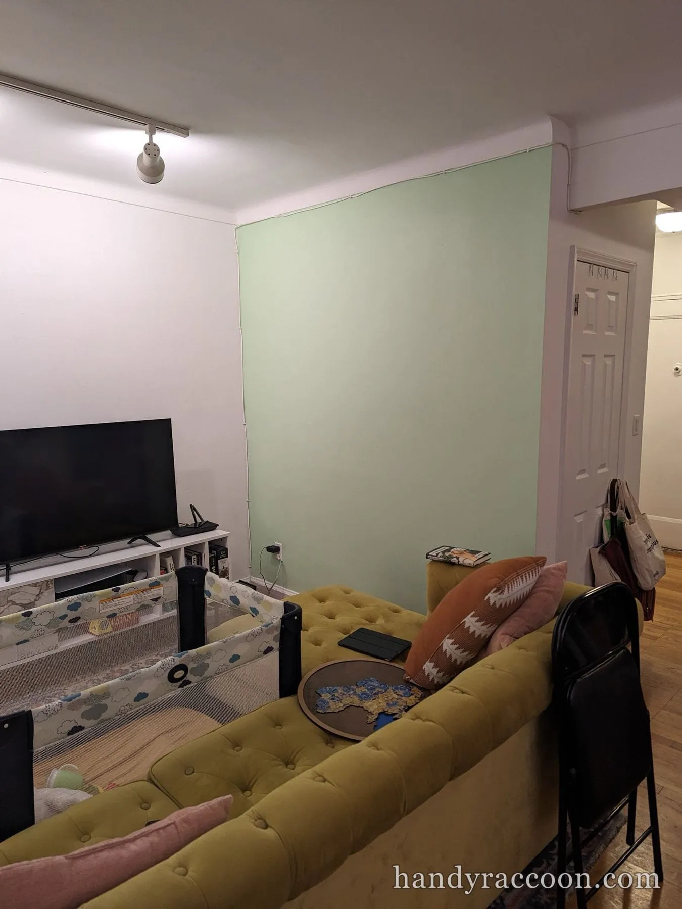 Living room with a freshly painted pale green accent wall next to a TV and yellow sofa.