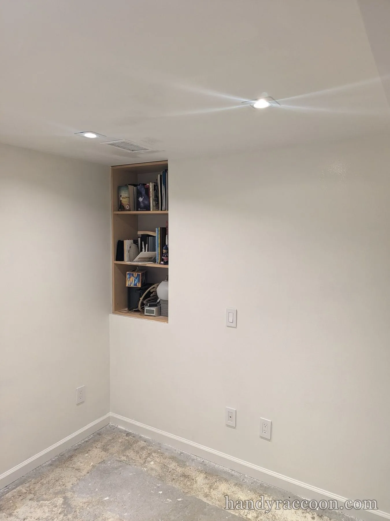 Room with freshly painted greige walls, a built-in bookshelf and bare concrete floor.