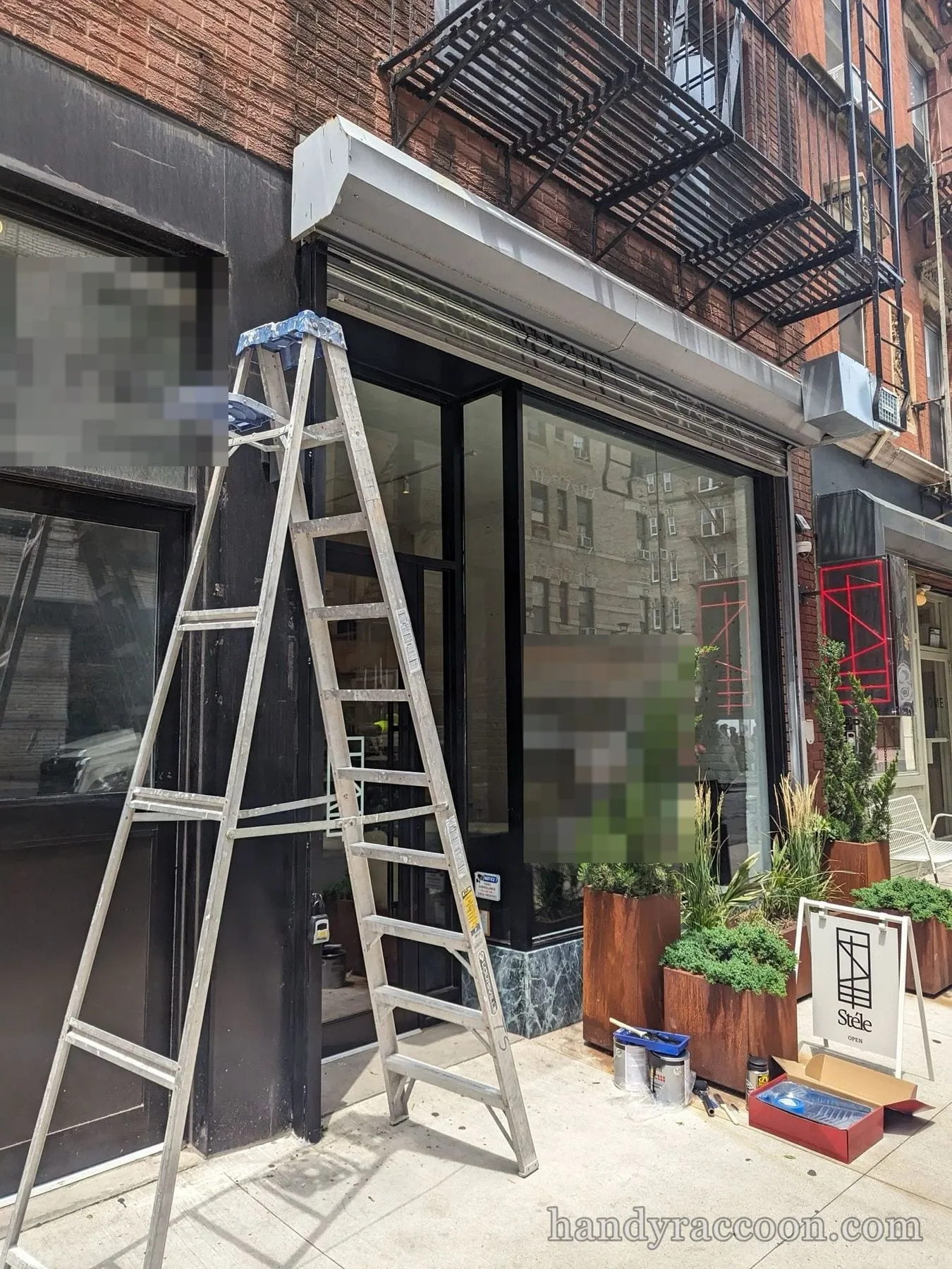 Painter's ladder in front of a black storefront being repainted, with a fire escape and brick building above.